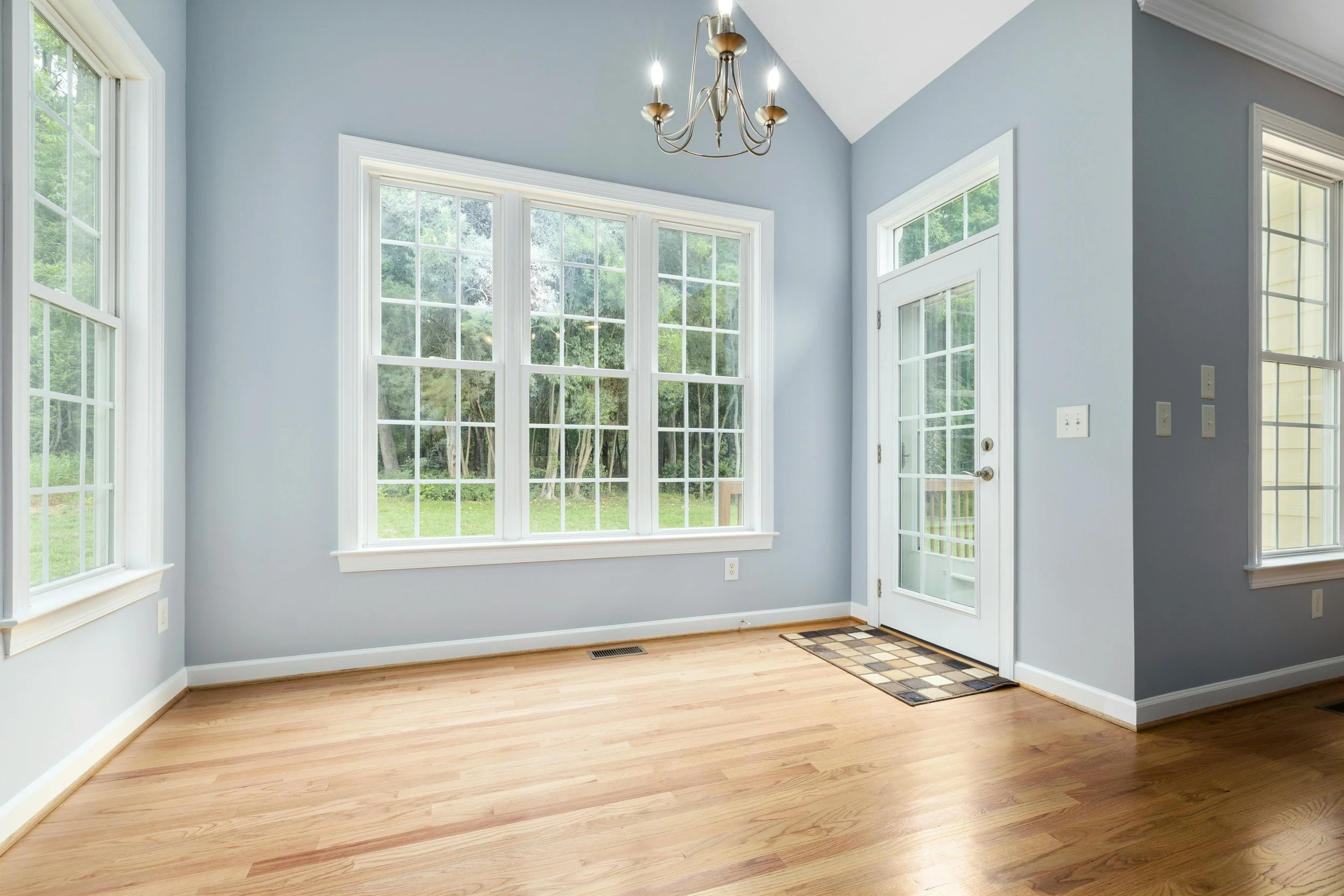 Empty room with large windows, light blue walls, wooden floors, a chandelier, and a glass door leading outside.