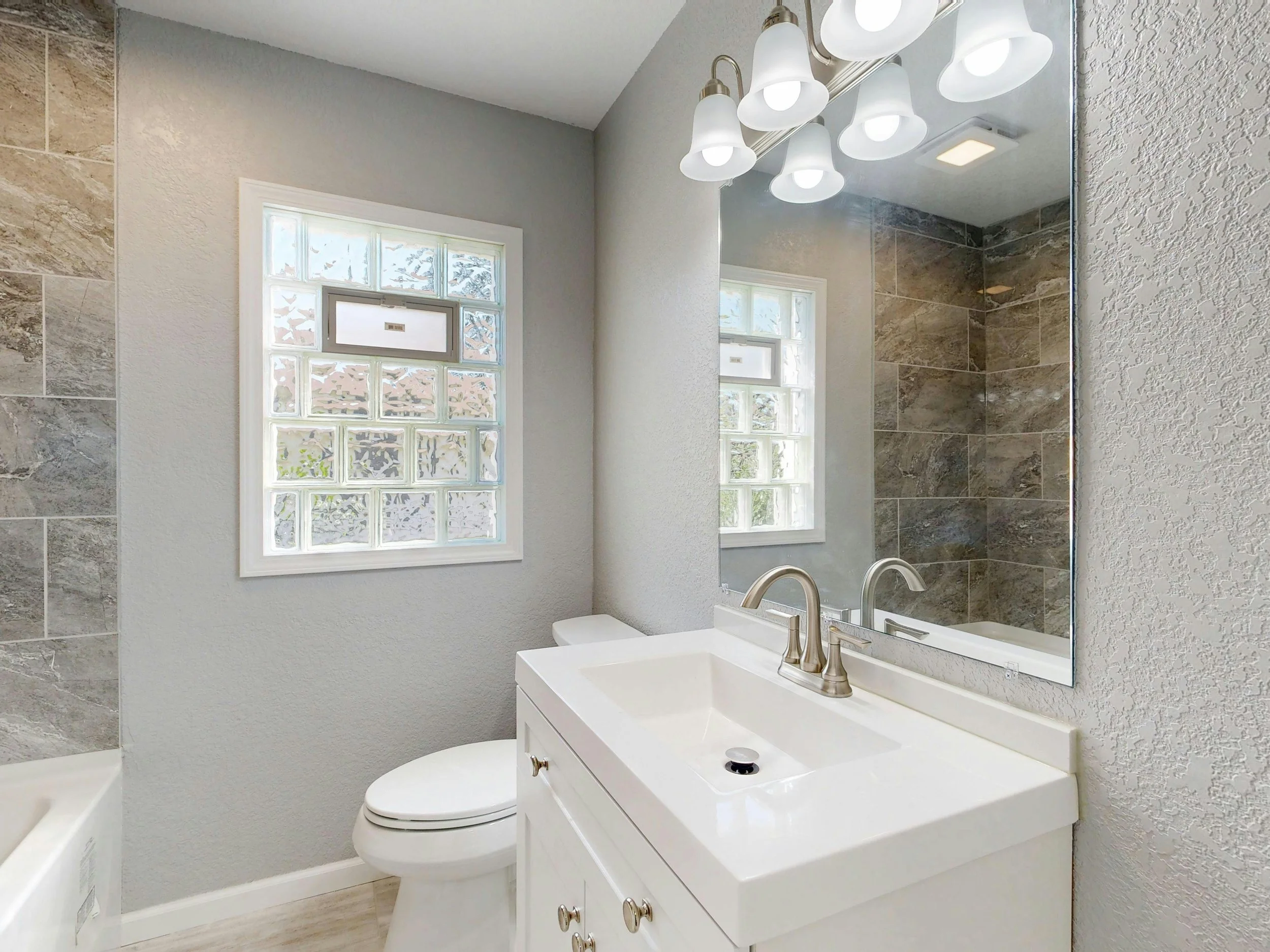 Bathroom with a white vanity, a sink with a chrome faucet, a large mirror, a wall light fixture with five bulbs, a frosted glass window, and a shower area with brown tile walls.