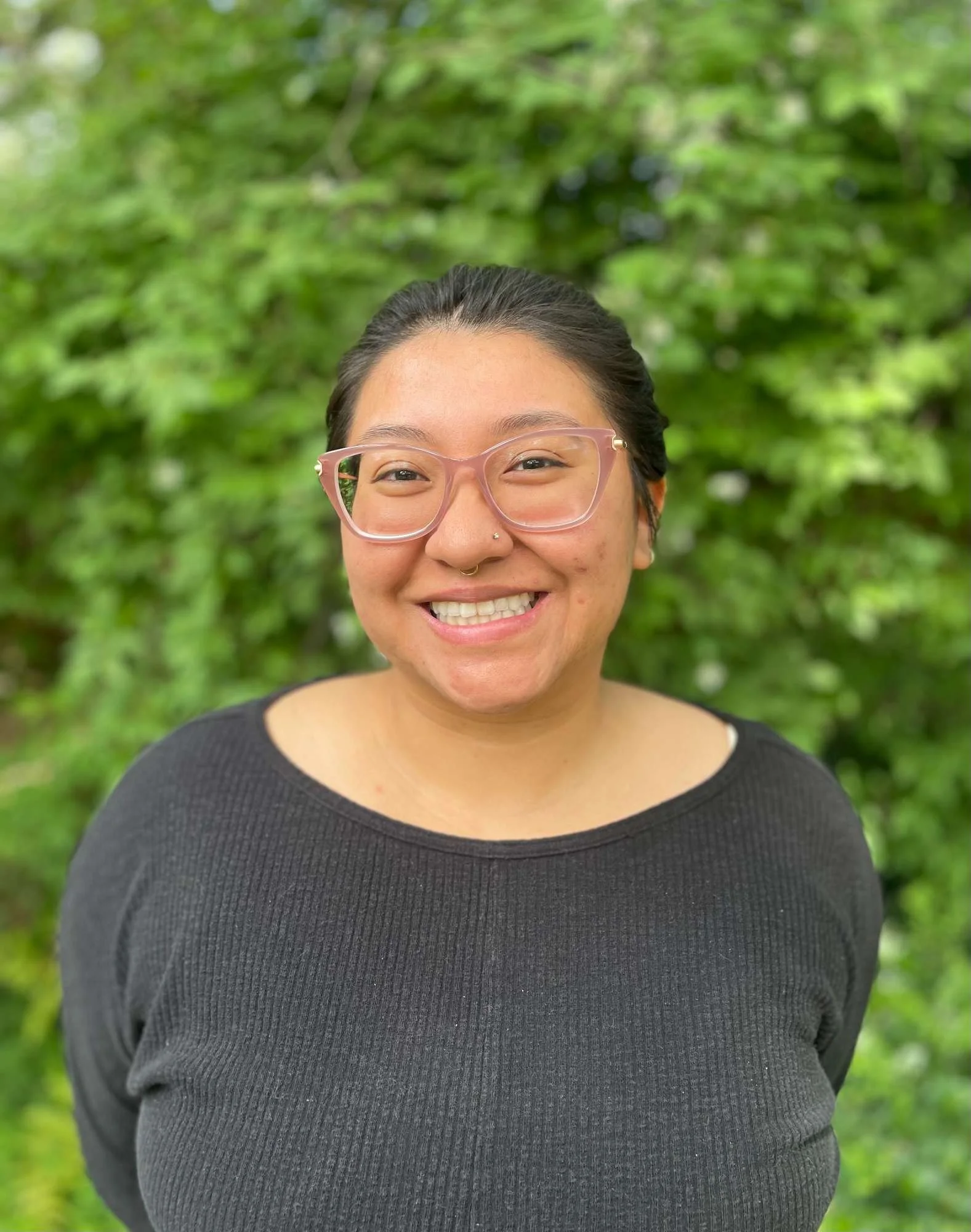 Lucia Mercado with black hair and glasses smiling outdoors with a green leafy background.