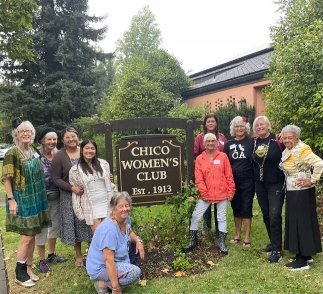 Group of women standing and sitting around a sign for the Chico Women's Club, established 1913, in a garden with trees and a building in the background.