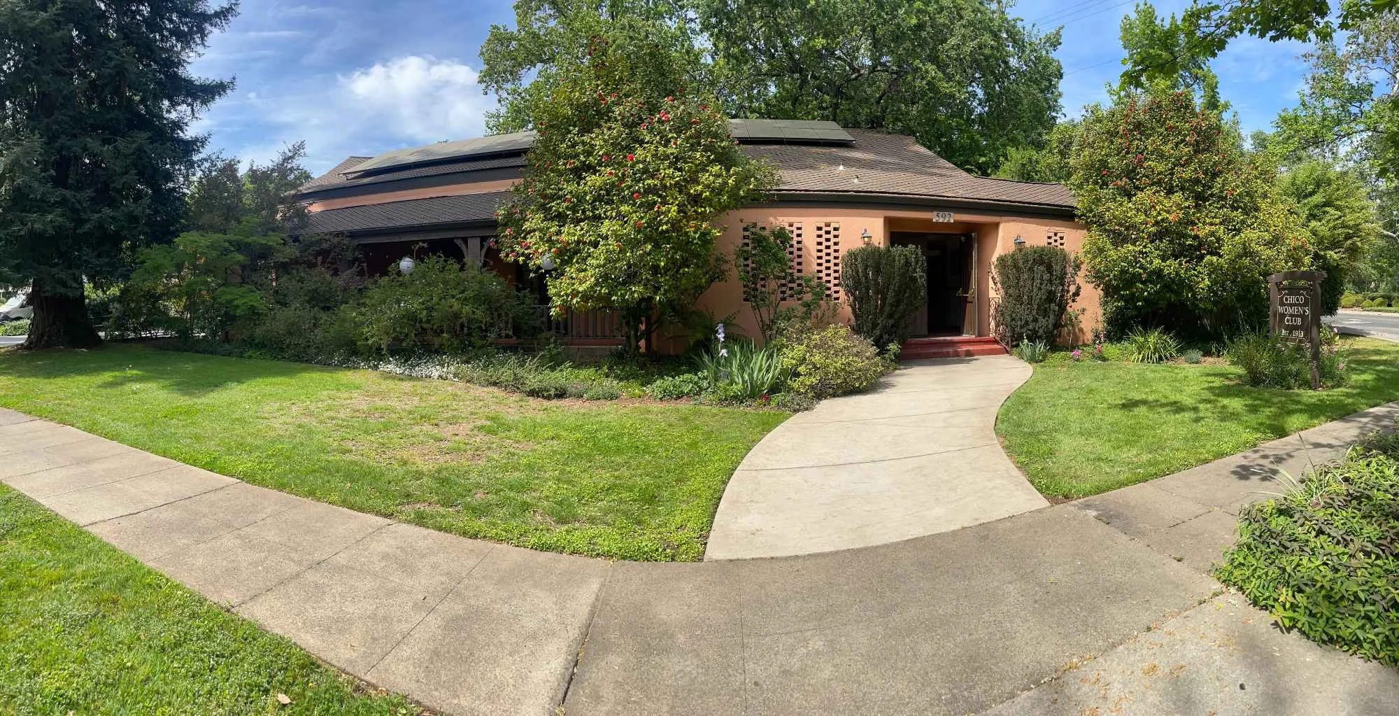 Chico Women's Clubhouse building with a curved roof, surrounded by green trees and bushes, with a concrete walkway leading to the entrance and a sign that reads 'Chico Women's Club'.