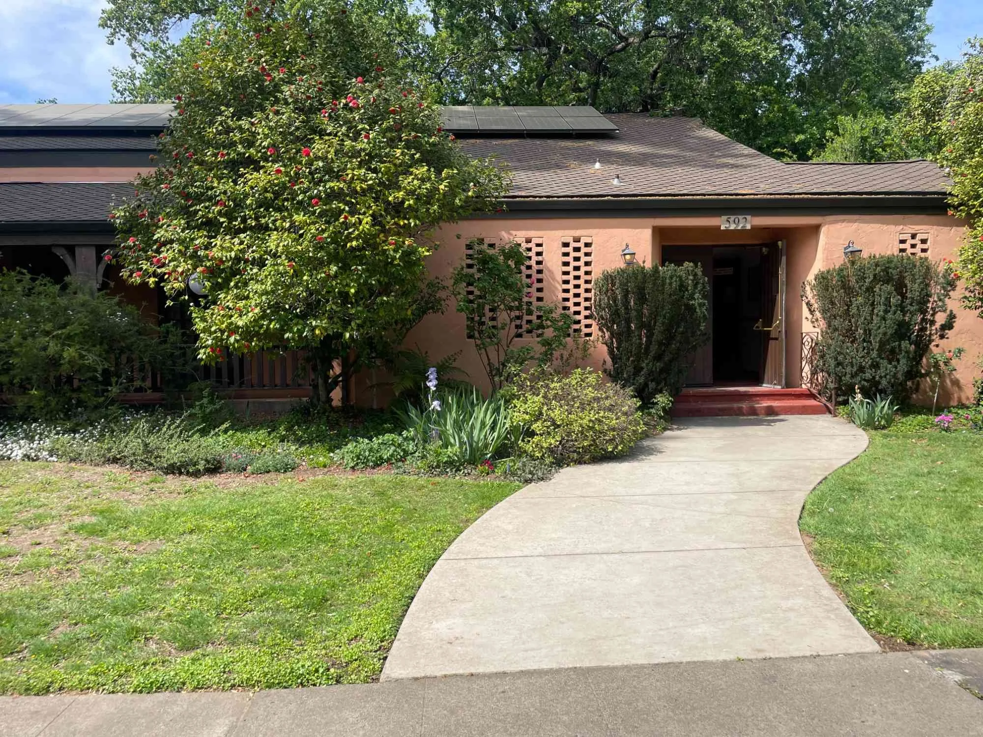 Chico Women's Clubhouse building with a curved walkway leading to the front door, surrounded by green bushes, trees, and flower beds under a partly cloudy sky.