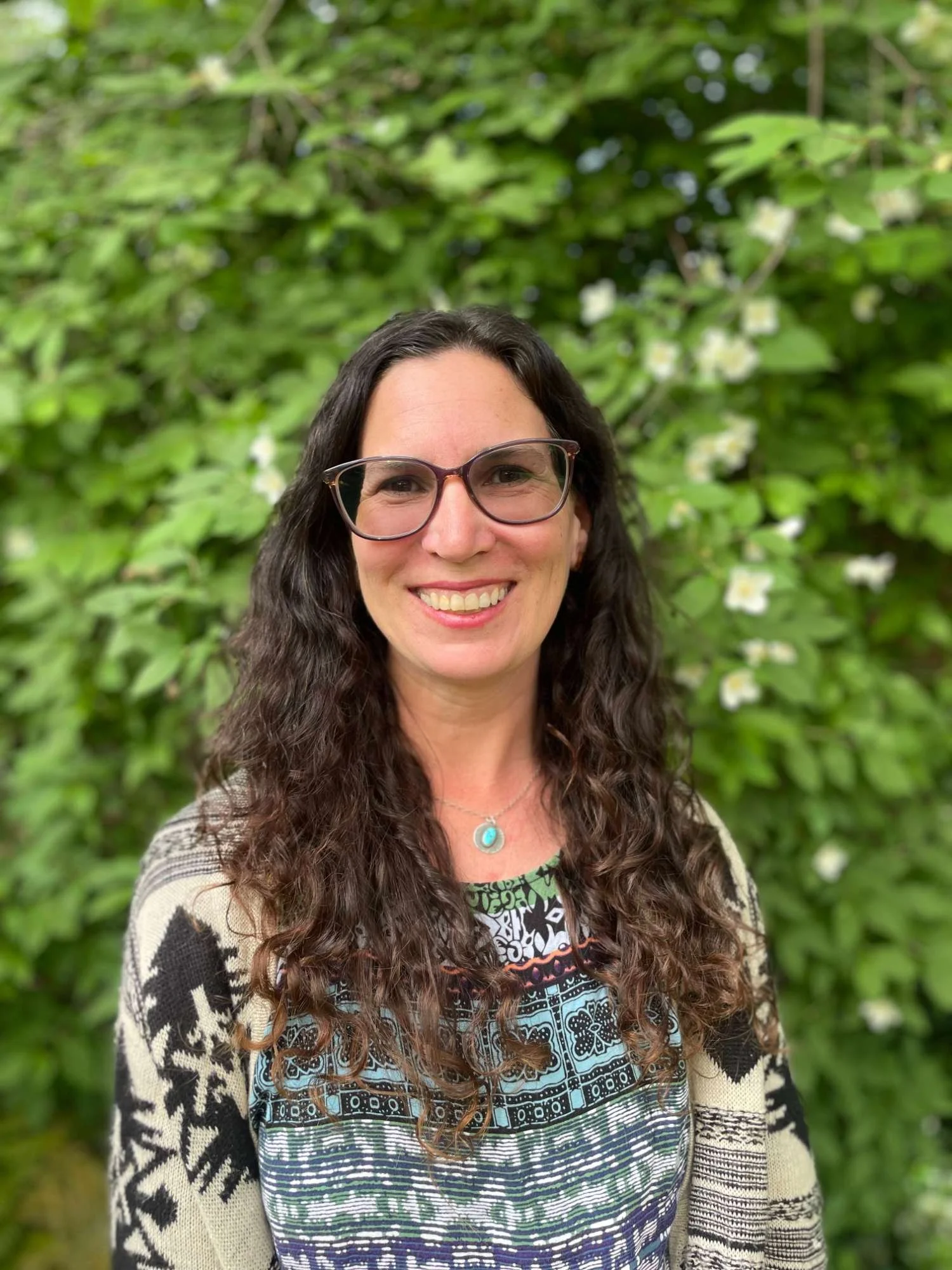 Morea Martin with long curly brown hair, wearing glasses and a colorful patterned top, standing in front of green foliage with white flowers.