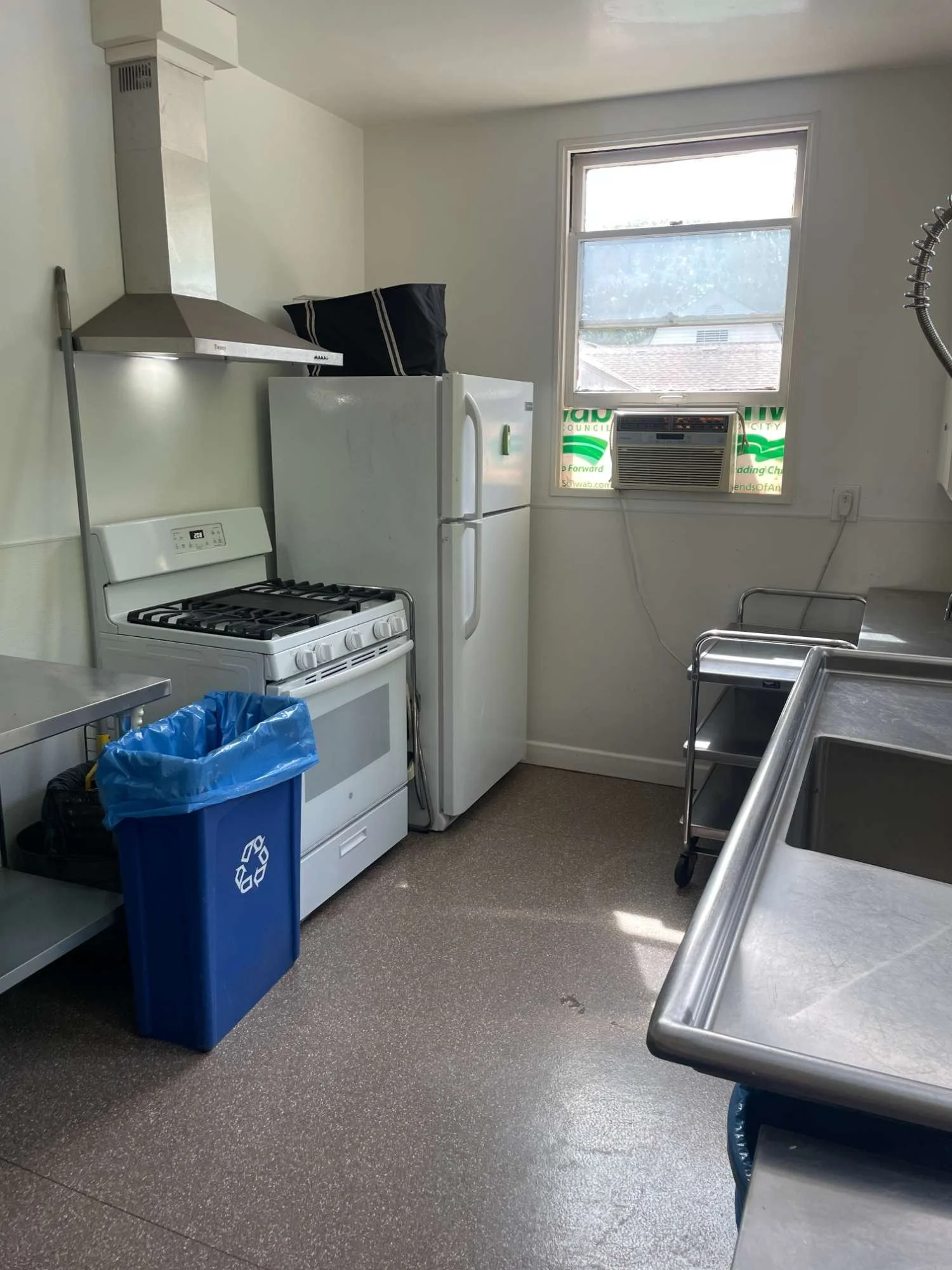 A small commercial kitchen in Chico Women's Clubhouse building with a window, a refrigerator, a stove, a trash can, and stainless steel sinks.