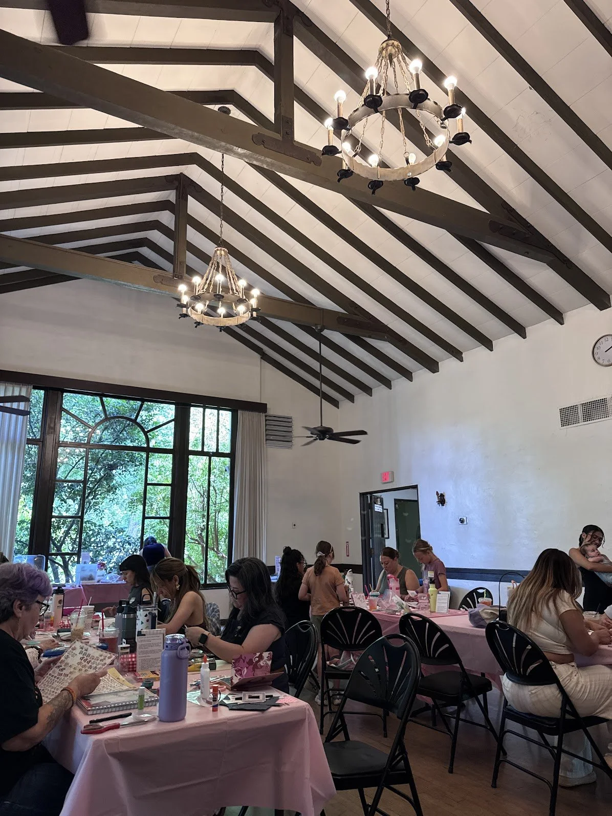 Inside the Chico Women's Club. People sitting at decorated tables in a bright room with large windows, chandeliers, and teal ceiling fans, engaging in a crafting event.