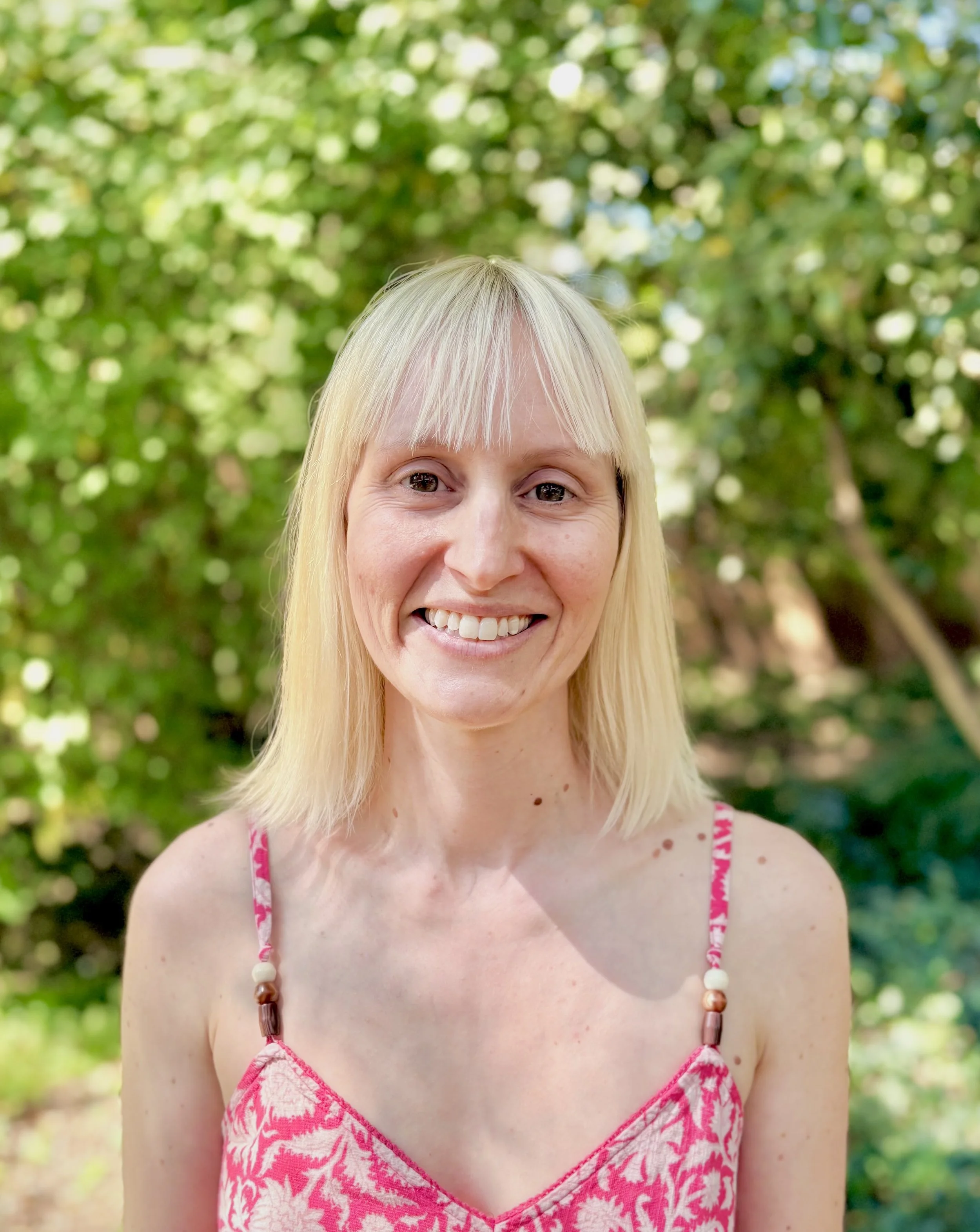 Alicia Trider with shoulder length blonde hair, wearing a strappy summer dress, stands outside in front of green foliage looking directly at the camera