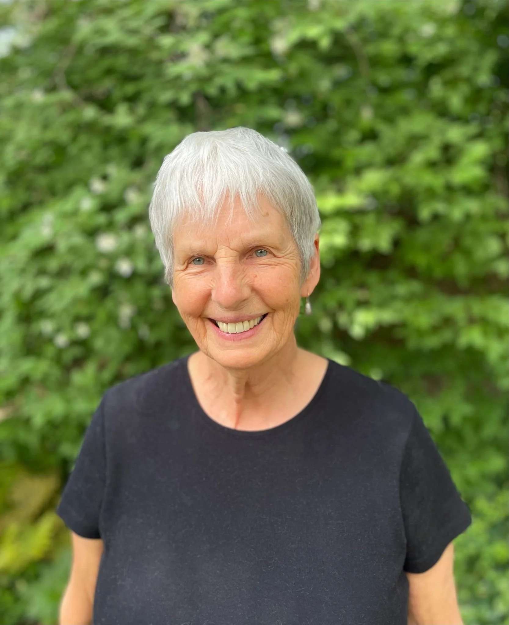 Joy Mendoza with short white hair smiling outdoors with green foliage in the background, wearing a black top.