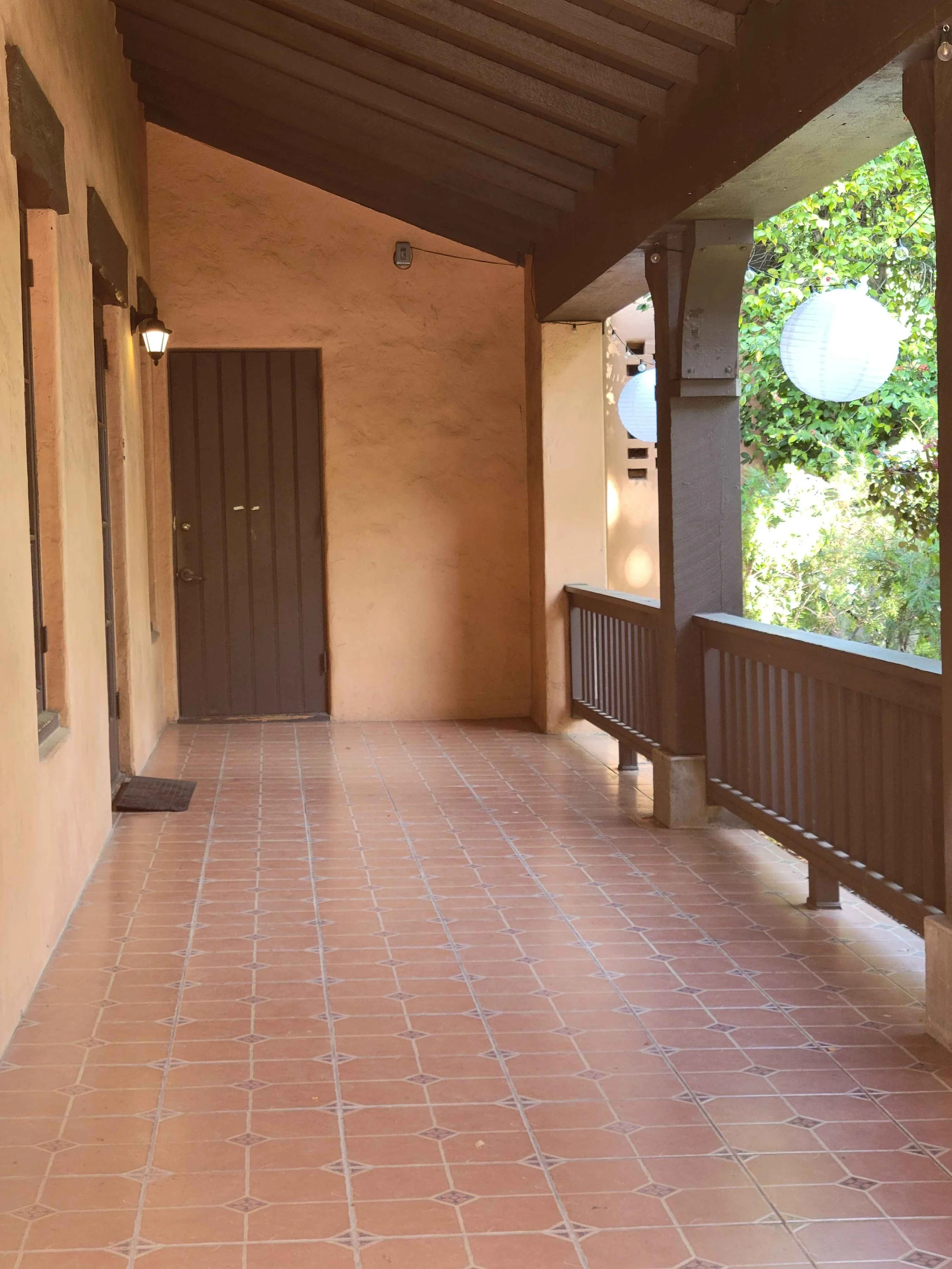 Chico Women's Clubhouse building outdoor porch with terracotta tile flooring, a brown door, a wooden railing, and paper lanterns hanging from the trees, with lush greenery in the background.