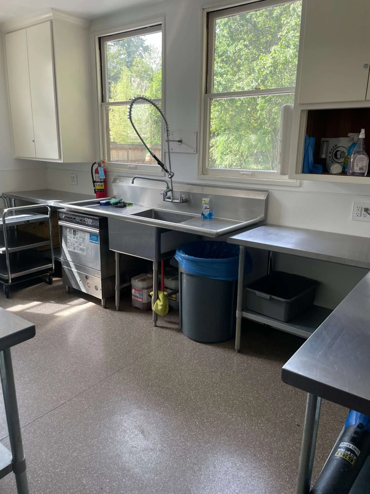 Commercial kitchen in Chico Women's Clubhouse building with stainless steel sink, windows, trash can, cleaning supplies, and stainless steel tables.