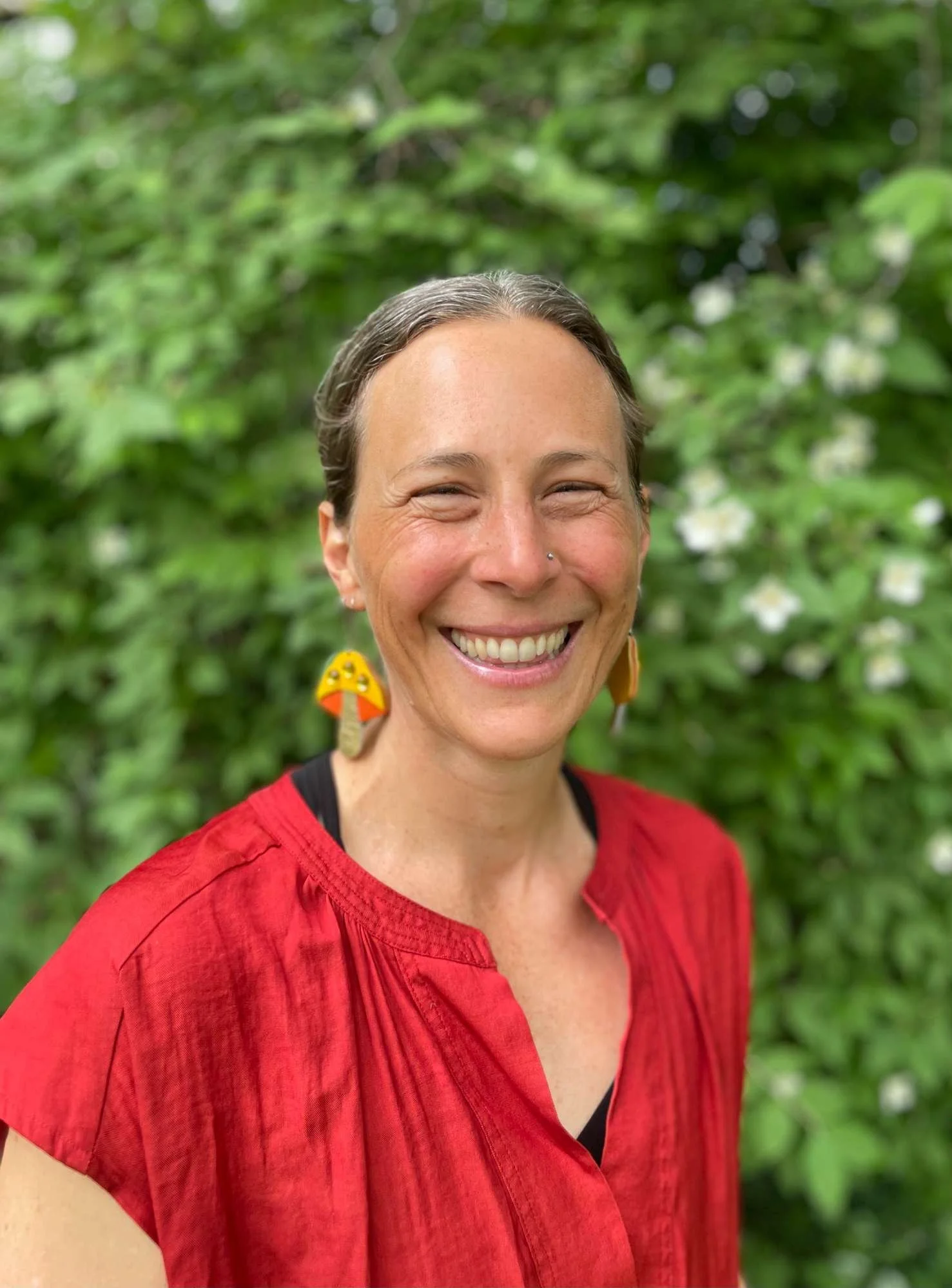Colleen Wofchuck smiling outdoors with a green leafy background, wearing red top and unique earrings.