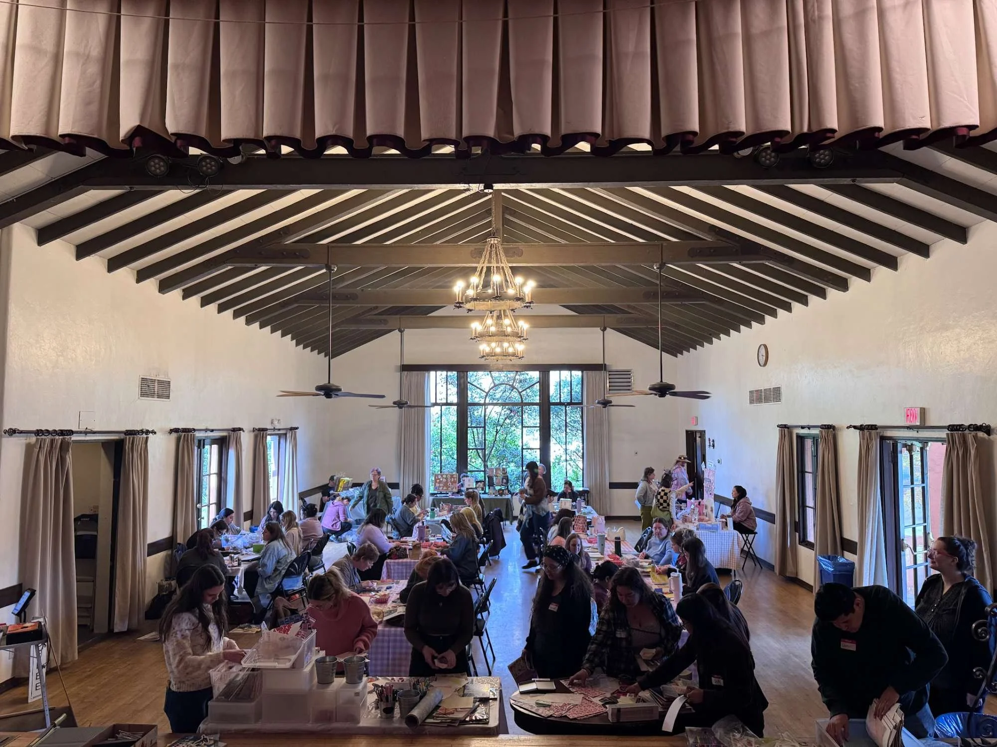 A large, bright room with high ceilings, chandeliers, and ceiling fans filled with people attending a craft or market event, with tables arranged along the walls and center, displaying various items.