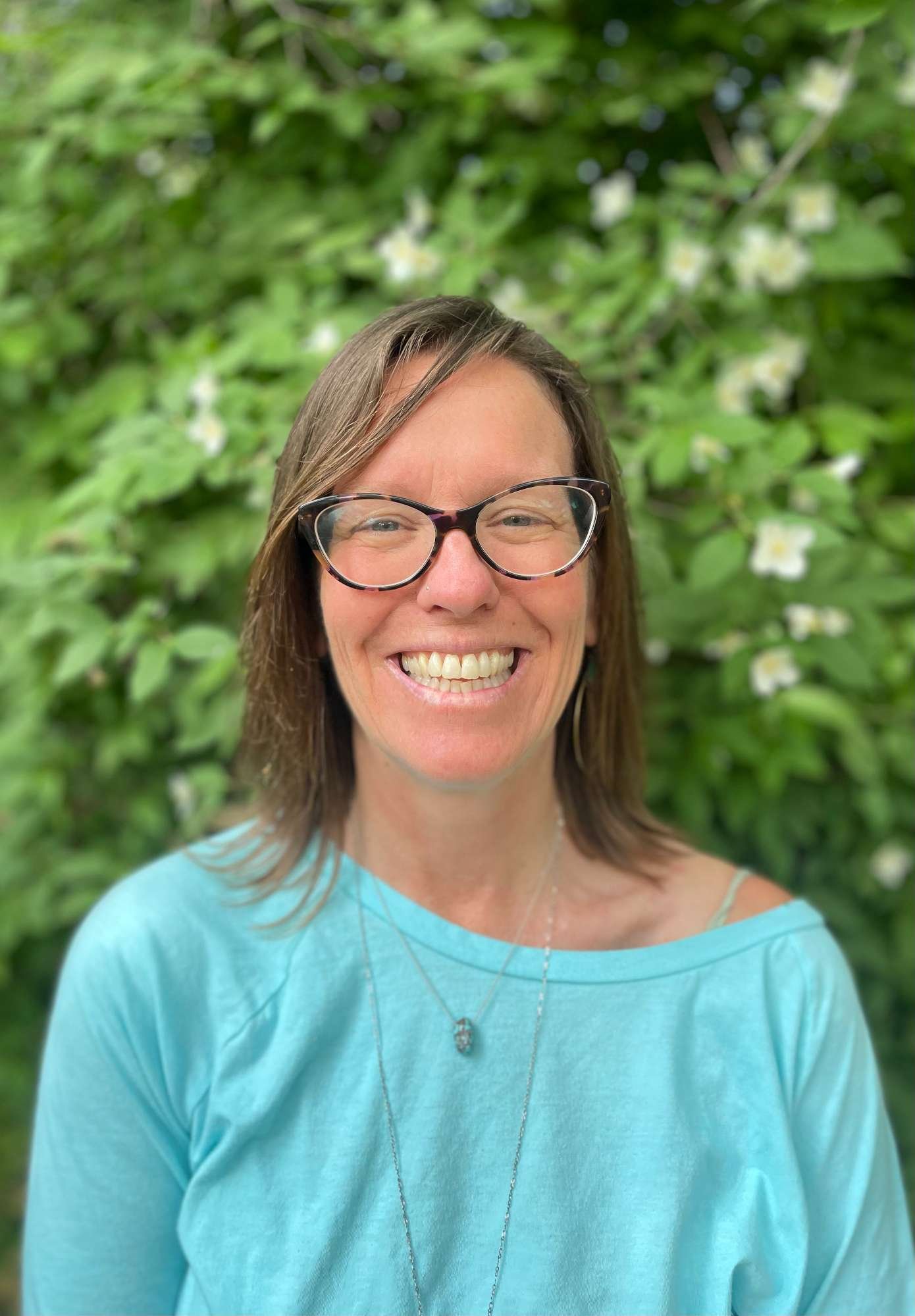 Serra Wells wearing glasses smiling outdoors in front of green foliage with white flowers.