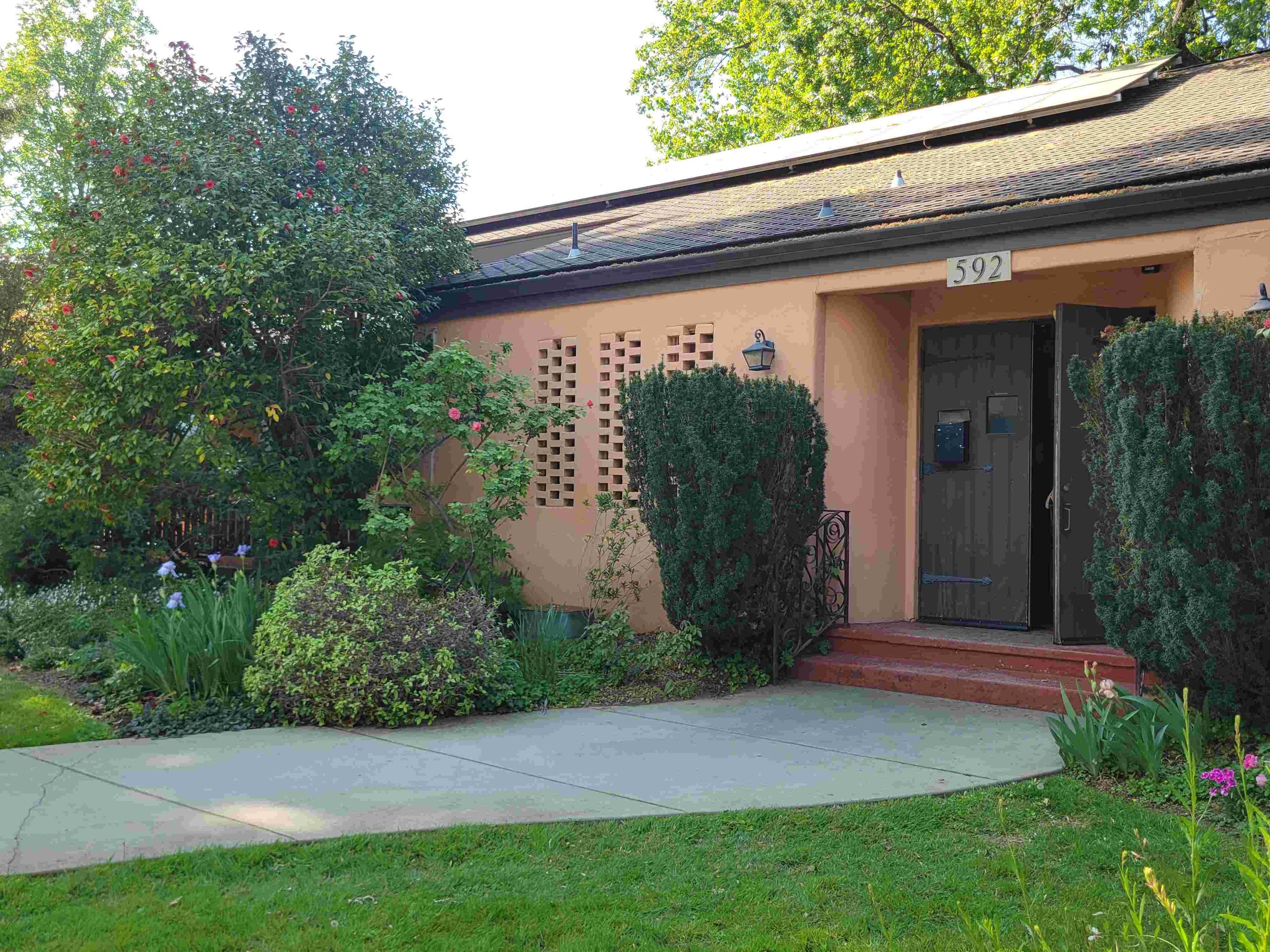 Chico Women's Club: front view of a house with pinkish stucco walls, a dark wooden door, and a black mailbox. There are plants and shrubs framing the walkway, a large tree with pink flowers on the left, and a green lawn in the foreground.