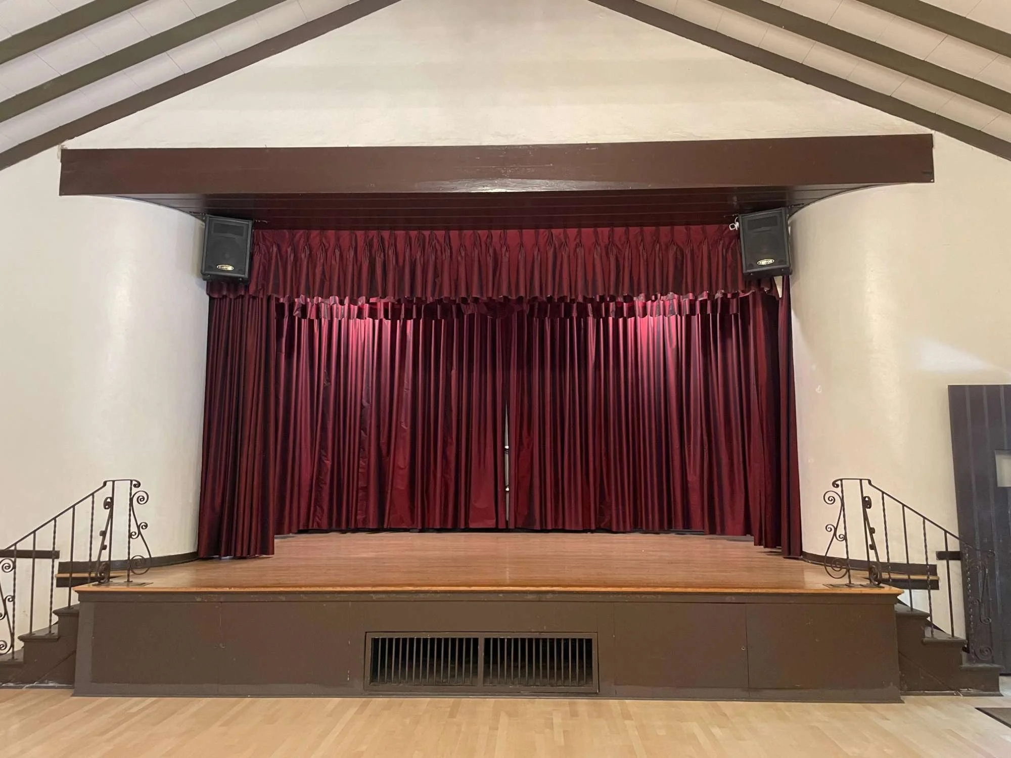 Empty stage in the chico Women's Club with closed red curtains, two speakers mounted on walls, and small stairs on either side.