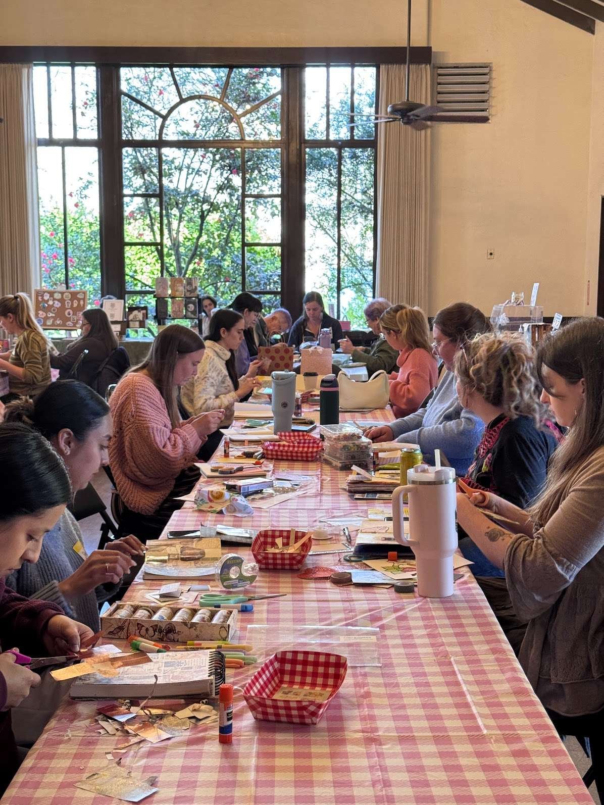 Inside the Chico Women's Club. People sitting at a long table with craft supplies, working on art projects, inside a room with large windows and a view of trees outside.