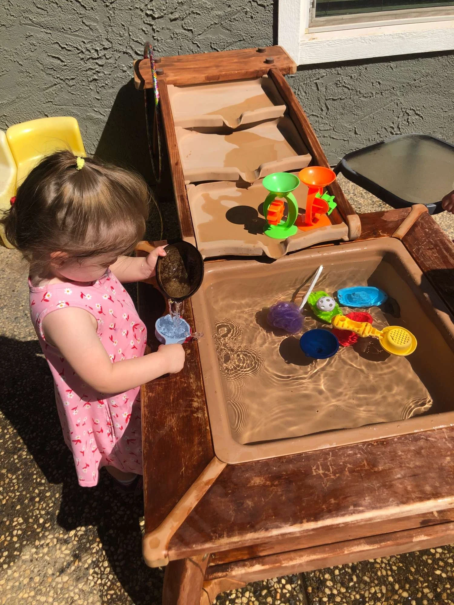 A young girl in a pink dress with flower patterns is playing at a water table, pouring water from a spoon into a cup. The water table has various toy boats and floating toys. The setting is outdoors near a gray wall and a window.