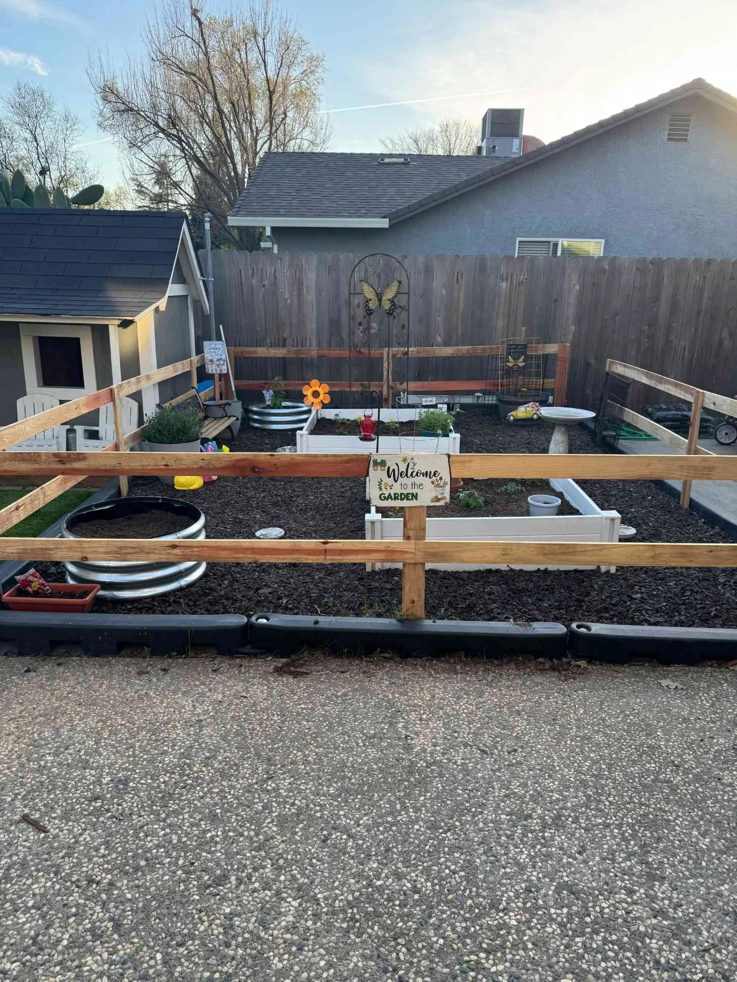 A small backyard garden enclosed by a wooden fence, with garden beds, a welcome sign, decorative butterfly, and outdoor toys.