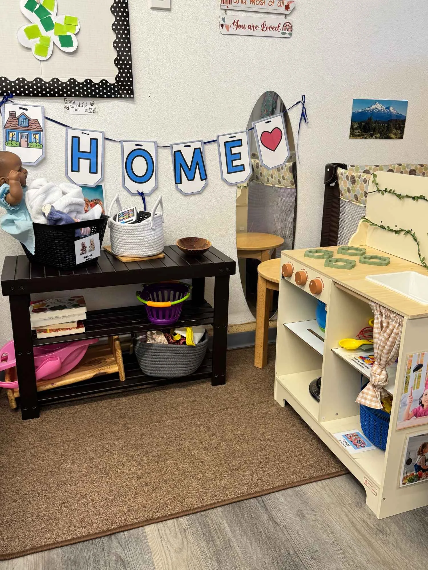 A classroom corner with a banner spelling 'HOME' decorated with a heart, a small play kitchen, a mirror, and various toys and books on shelves.