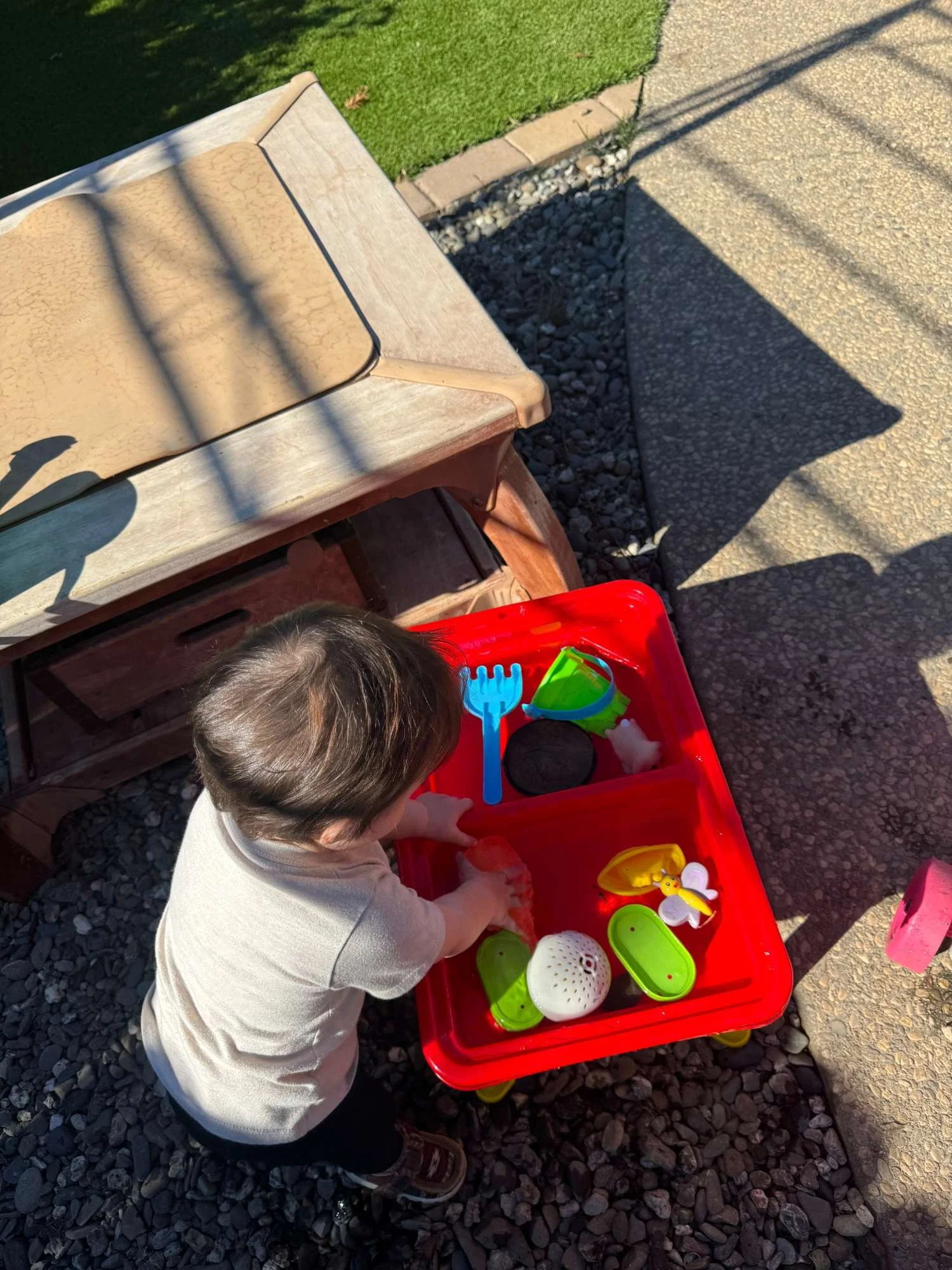 A young child playing with a toy bathroom set in a red plastic tub outdoors on a gravel surface.
