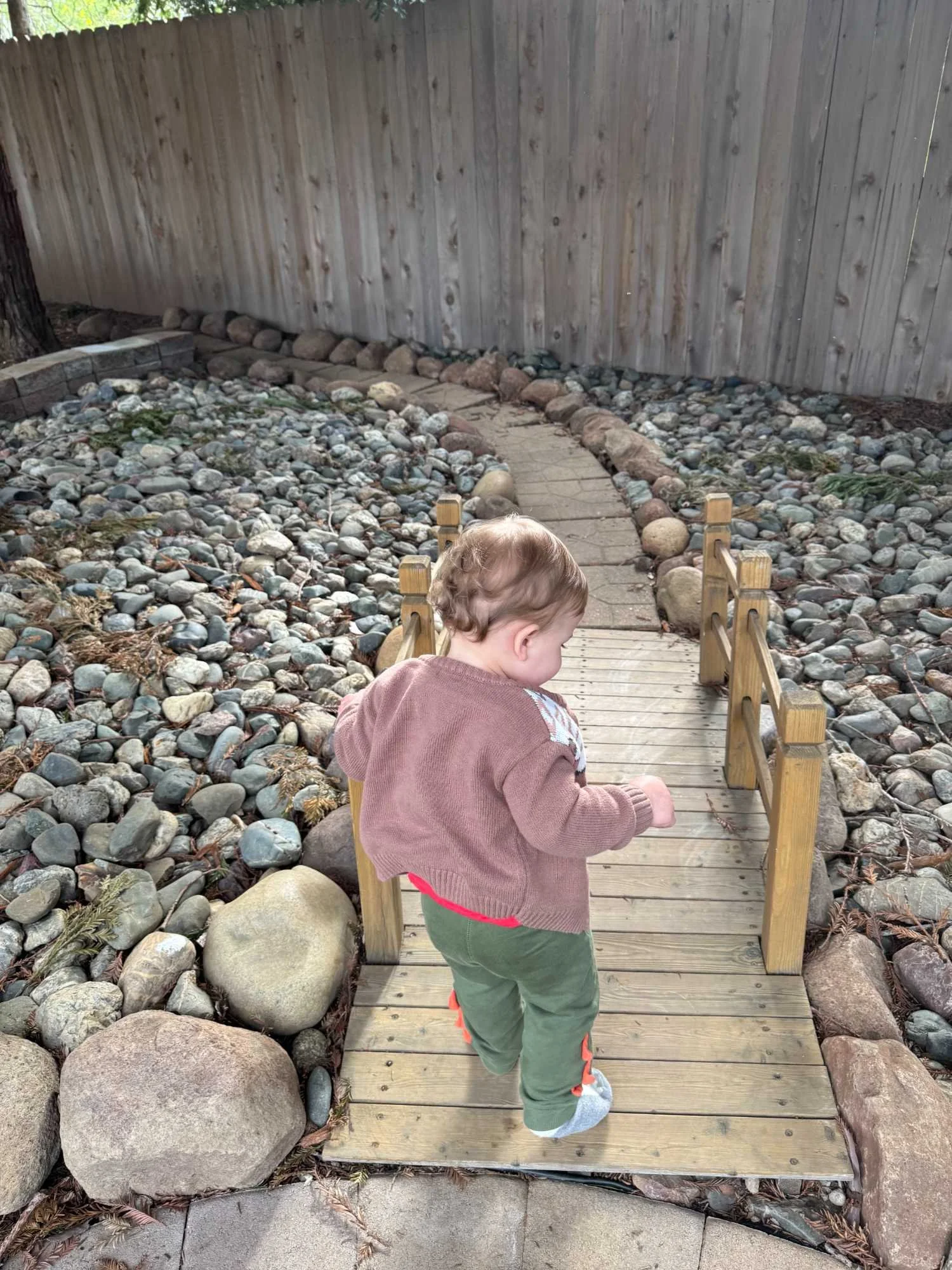 A young child walking on a small wooden footbridge over a rocky yard, with a wooden fence in the background.