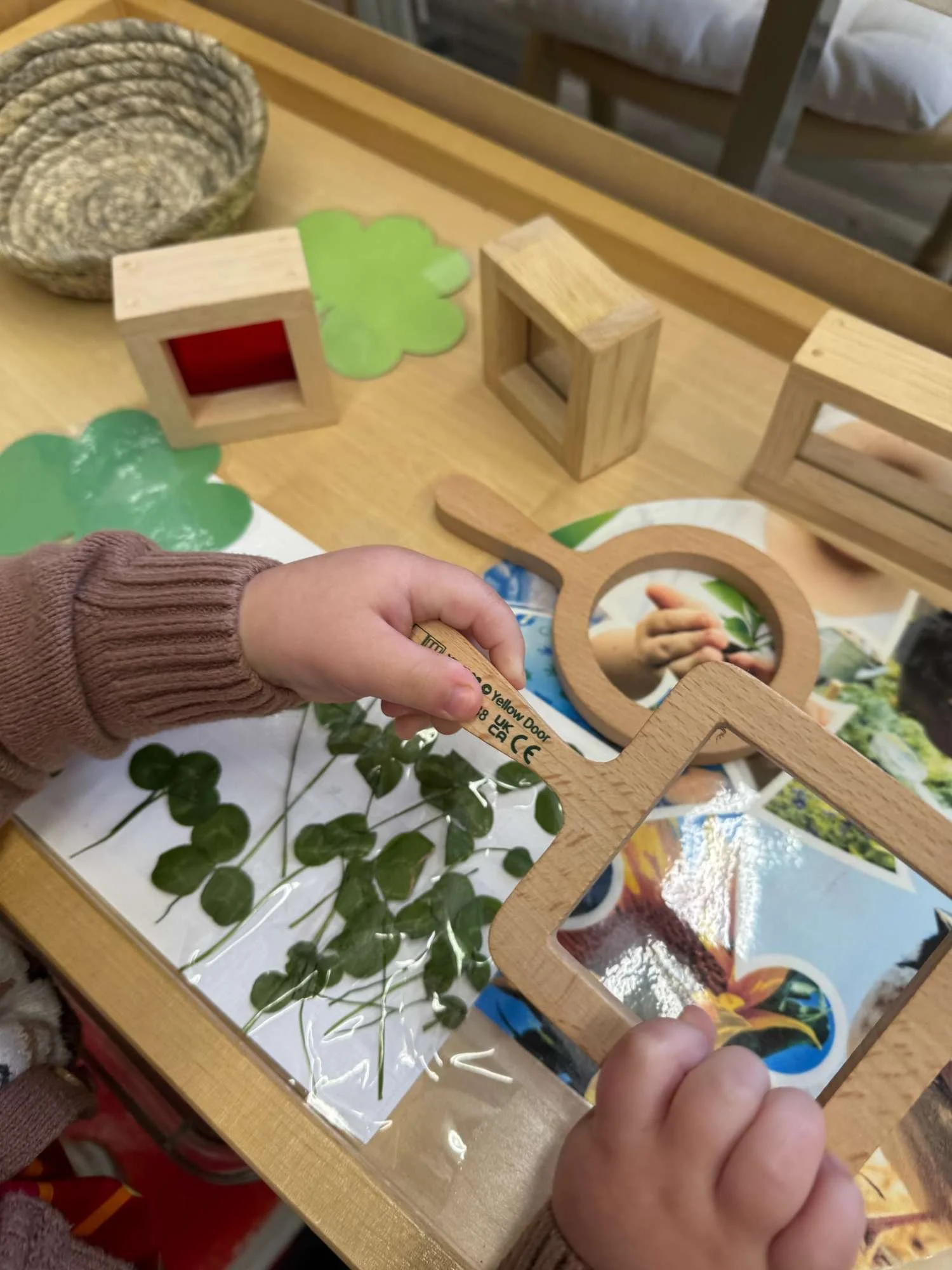 Child holding a wooden frame mirror while exploring a plant with green leaves on a clear sheet, on a wooden table with toy blocks, a wicker basket, and a mirror in the background.