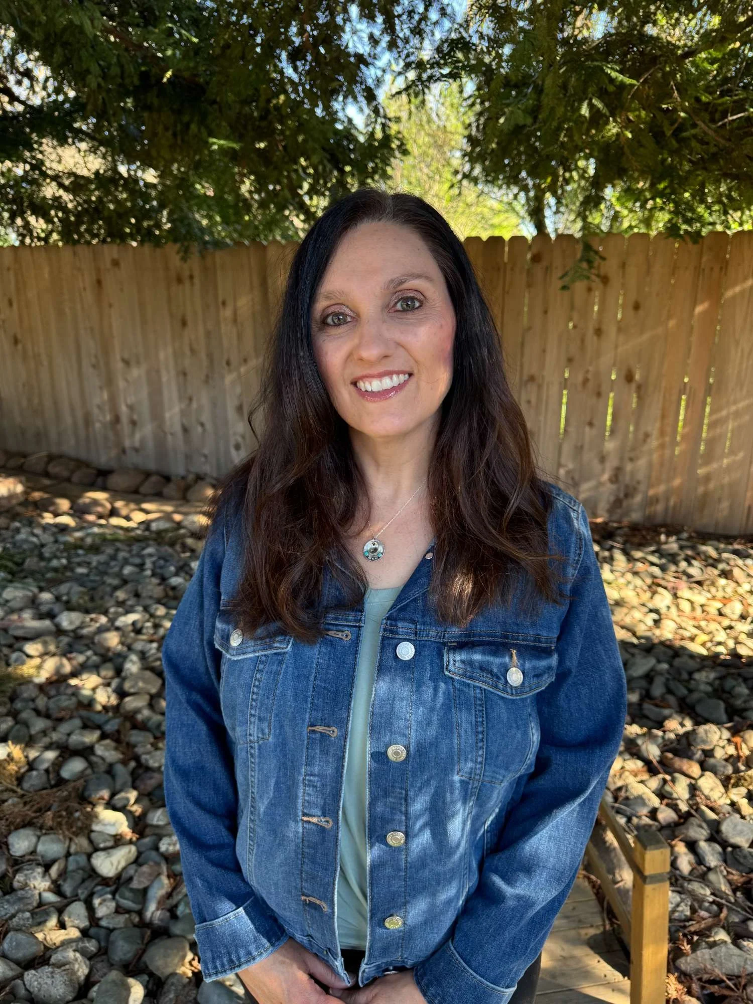 Little Hands owner, Suzi Maloney, smiling outdoors in front of a wooden fence and green trees, wearing a denim jacket and a necklace.