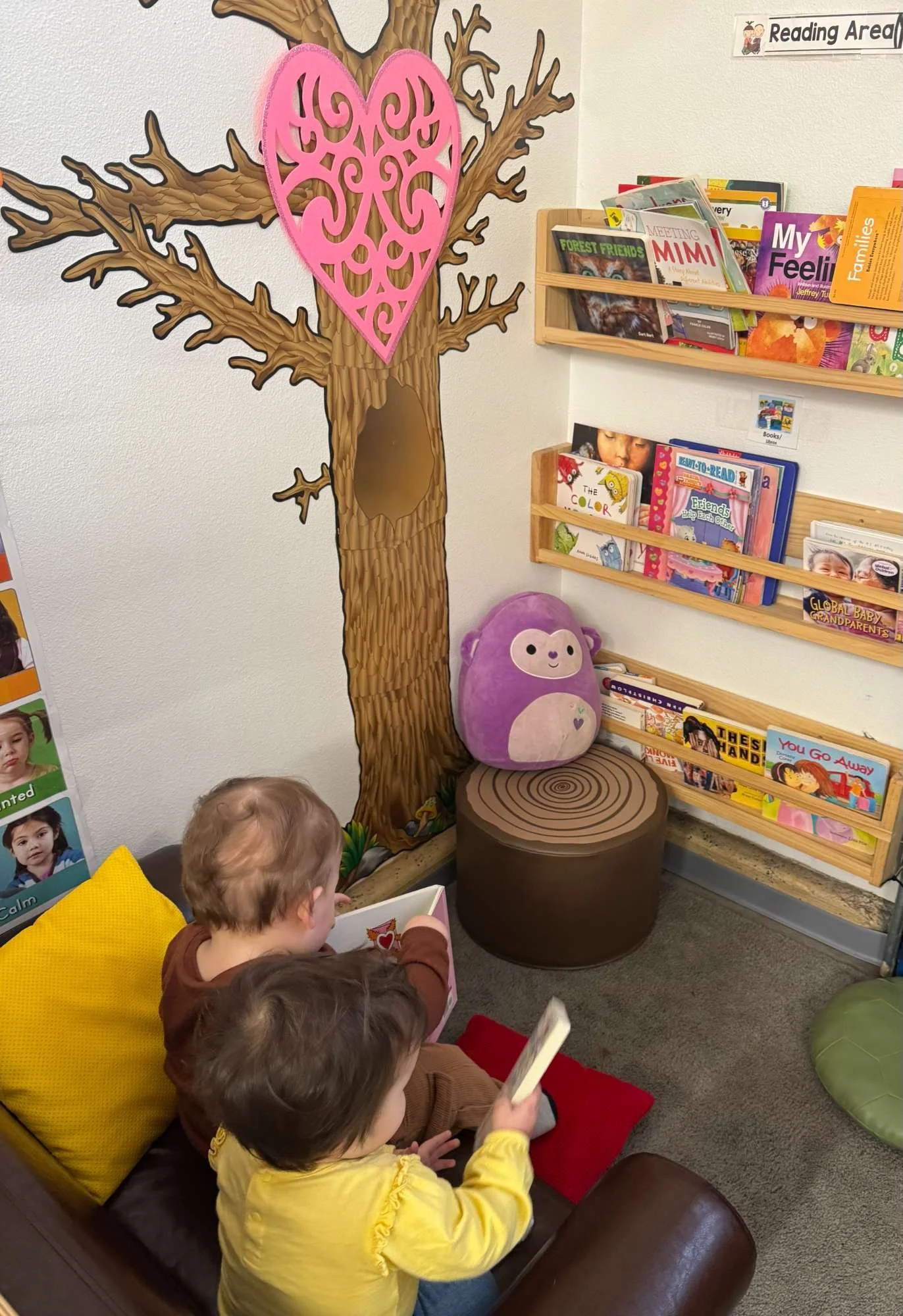 Children sitting on a brown couch in a children's reading area, with a decorative tree wall and a pink heart-shaped decoration, bookshelf filled with children's books, and a plush purple monkey pillow.