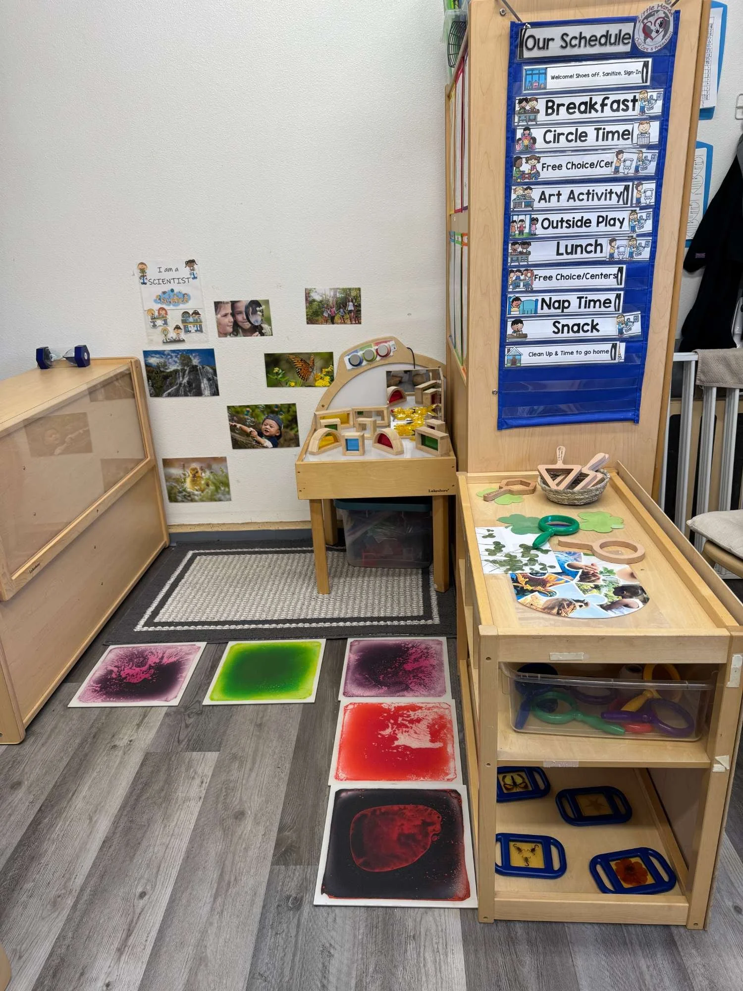 Classroom play area with educational materials, artwork, photographs, a play table, and a weekly schedule on a blue pocket chart. The schedule includes activities like breakfast, circle time, art, outside play, lunch, nap, and snack.