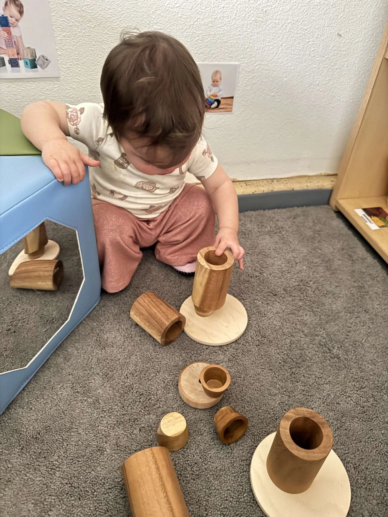 A young child with brown hair, wearing a white shirt with animal prints and pink pants, is playing with wooden stacking toys on a gray carpeted floor. The toys include various cylindrical and rounded pieces with round bases.