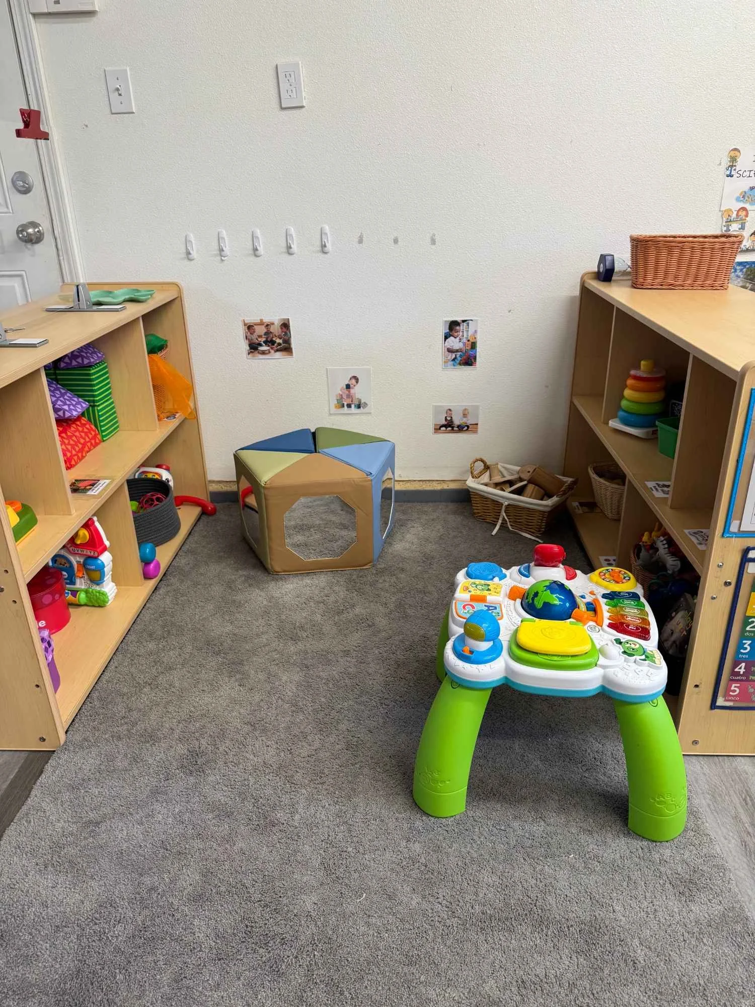 A children's playroom with wooden shelving units filled with toys and books, a colorful activity table, and pictures on the wall.