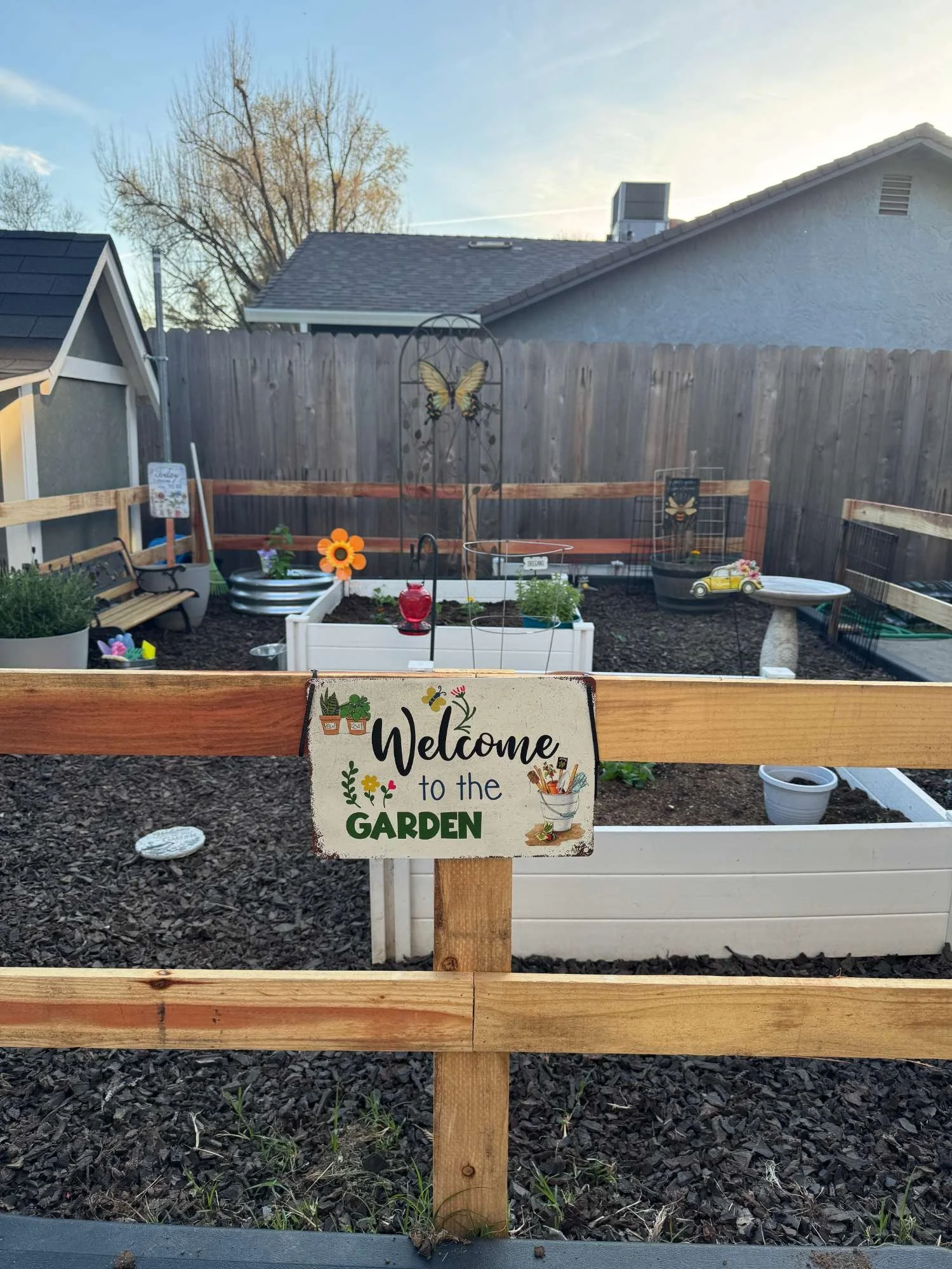 A small backyard garden with a wooden fence, raised garden beds, and decorative items. A sign on the fence reads 'Welcome to the Garden' with colorful gardening illustrations.