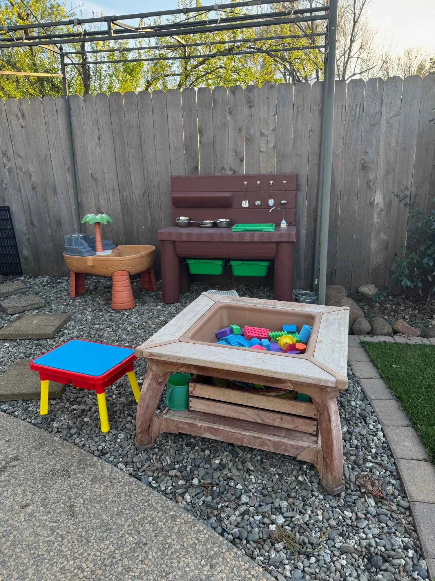 Children's outdoor play area with sandbox, play kitchen, small table, and toy set surrounded by gravel, pavers, and a wooden fence.