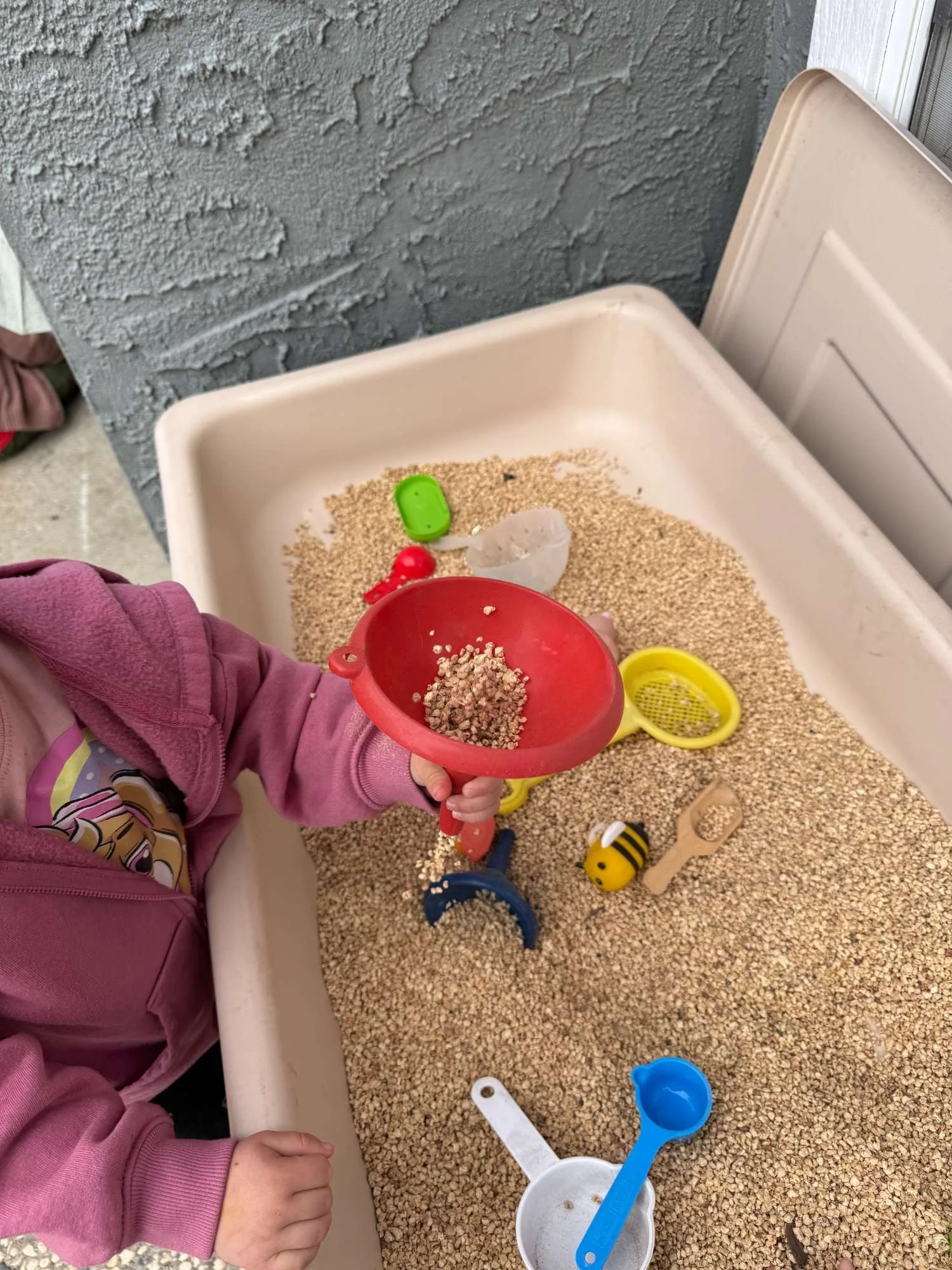 Child playing with sand and toy kitchen utensils in a sandbox, holding a red scoop filled with sand, surrounded by various toys including a yellow spoon, a small bee toy, and a blue shovel.