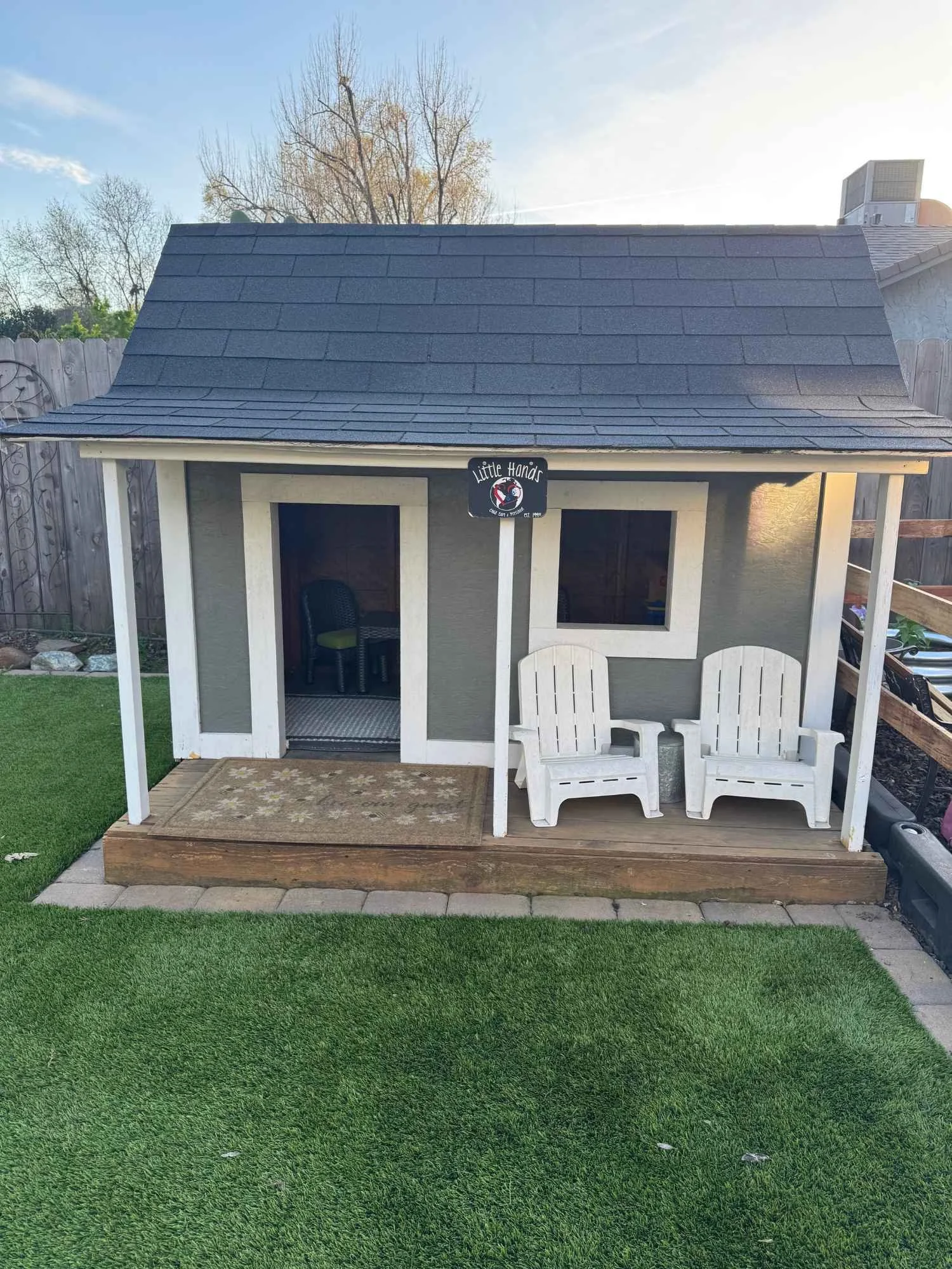 A small outdoor playhouse with a blue shingle roof, gray walls with white trim, two white Adirondack chairs on a wooden deck, and a sign that reads 'Little Hands' hanging from a post. The playhouse has a doorway with an interior visible and a window.