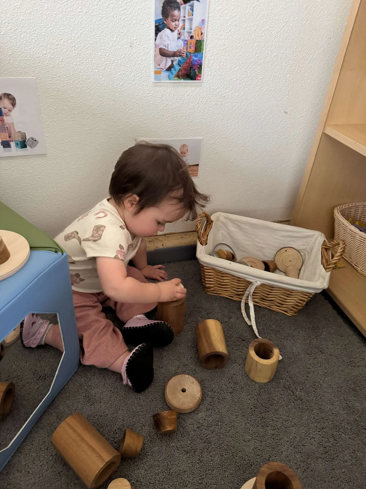 A young child with short brown hair, wearing a cream shirt with patterns and pink pants, is playing with wooden cylindrical blocks on a dark gray carpeted floor in a playroom. There is a blue table to the left and a wicker basket filled with more woo