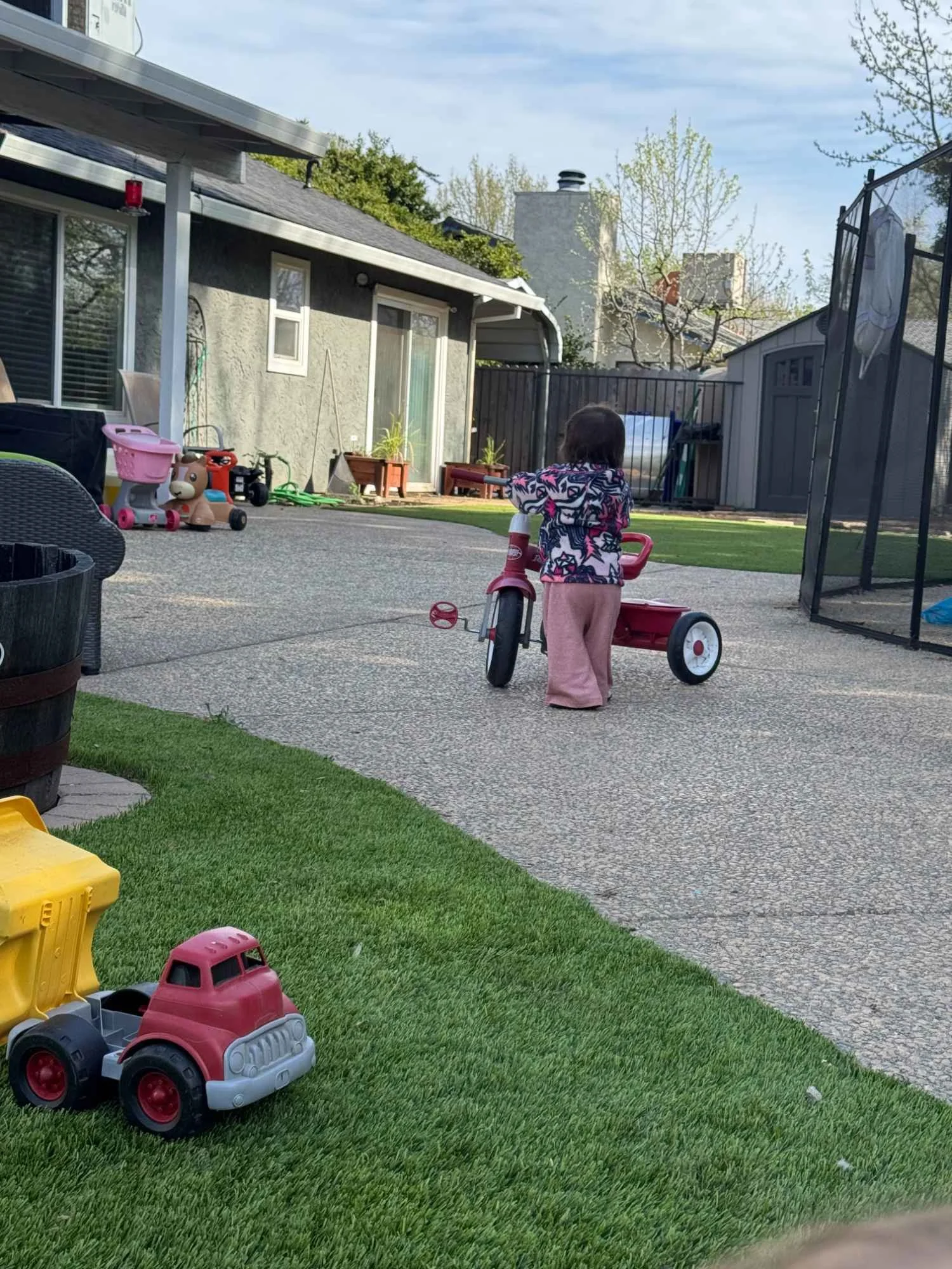Child playing outside in a backyard with toy vehicles, including a red and gray toy truck in the foreground and a tricycle nearby, on a mix of grass and concrete patio with house and garden items in the background.