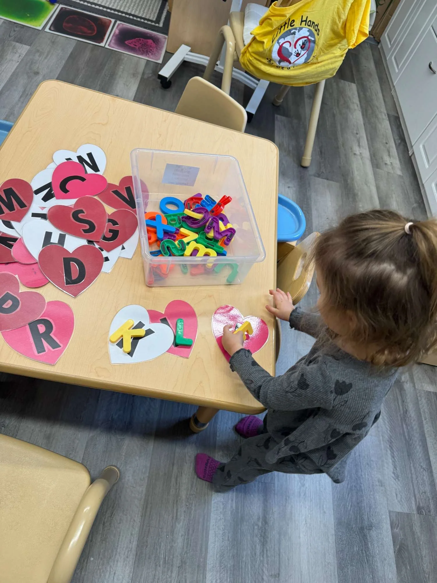 Little girl placing magnetic letter on a heart-shaped paper in a classroom. The table has magnetic letters and heart-shaped papers with letters. There are chairs, classroom cabinets, and educational materials in the background.