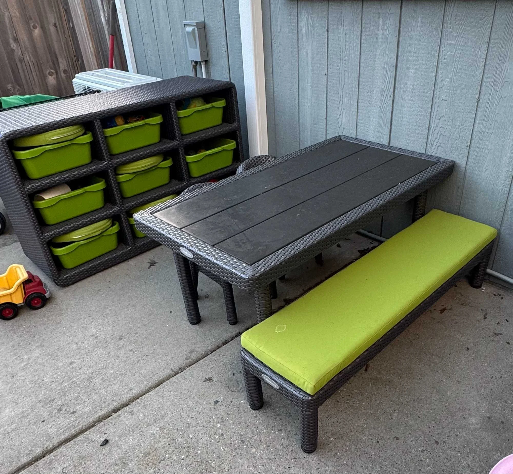 Outdoor patio with a black wicker table, a matching bench with a yellow cushion, and a storage unit with multiple green outdoor storage bins.