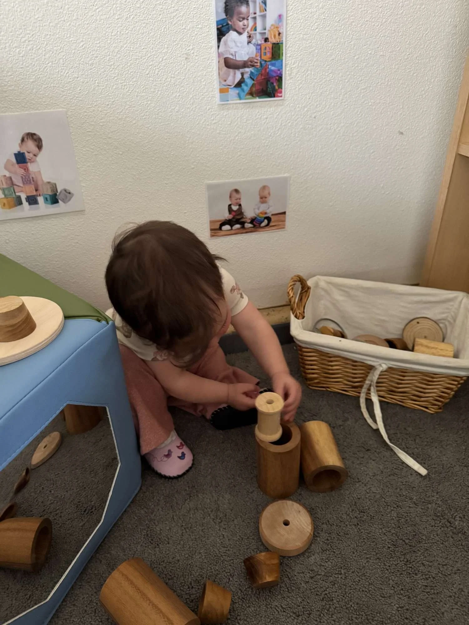 A young child playing with wooden blocks on the floor in a playroom, with a basket of more wooden toys nearby and photos of children on the wall.
