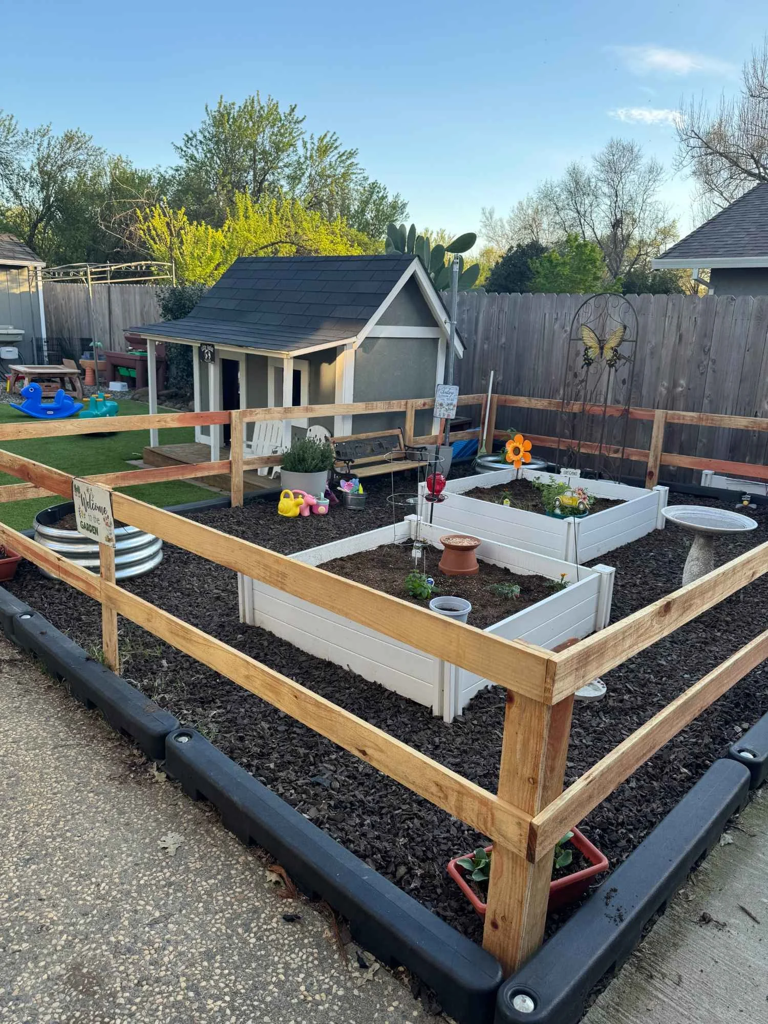 A backyard garden with raised white garden beds, a small doghouse, and a wooden fence. Children’s toys including a sandbox, watering cans, and paddling pool are visible. Trees and blue sky are in the background.
