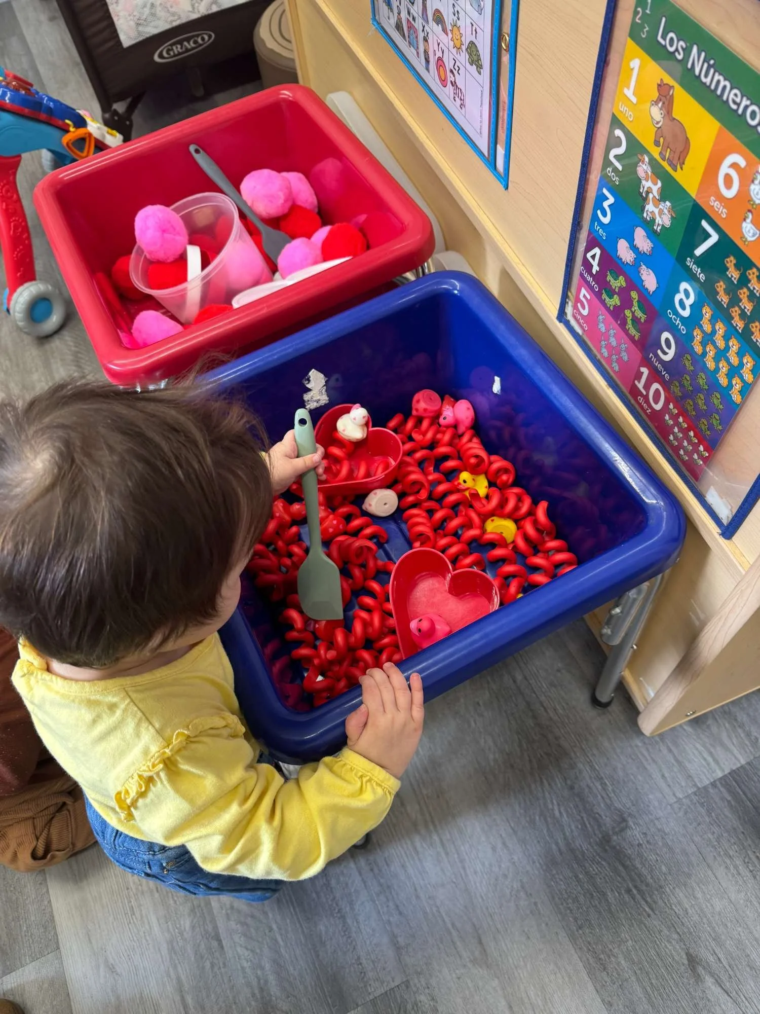 A young child playing with red and pink plastic food toys in a large blue toy bin at a preschool or daycare.