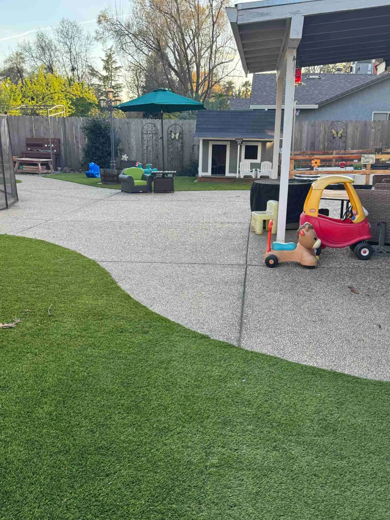 Child's backyard with a playhouse, toy car, and outdoor furniture on a concrete patio, with a grassy area in the foreground and trees in the background.