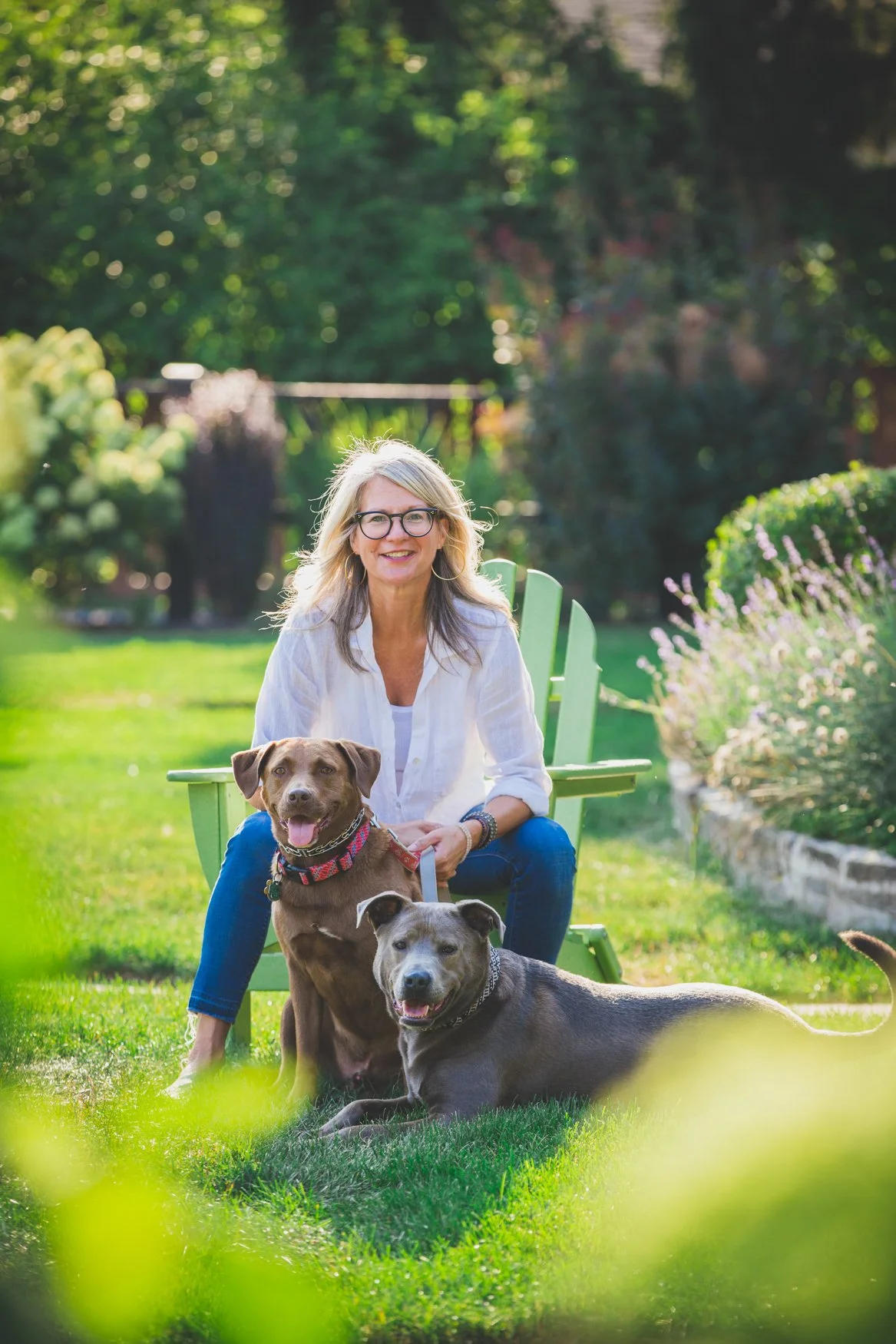 A woman with gray hair, glasses, and a white blouse sitting outdoors on a green bench, smiling with two dogs on the grass in front of her, one sitting and one lying down, in a lush garden with trees and flowering plants.