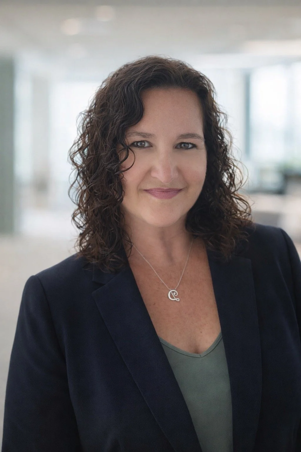 A professional woman with curly brown hair, wearing a dark blazer and a necklace with a pendant, smiling in a modern office environment.