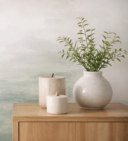 A wooden table holding three white candles of different sizes and a white textured vase with green leafy plants inside, set against a pale gradient wall background.