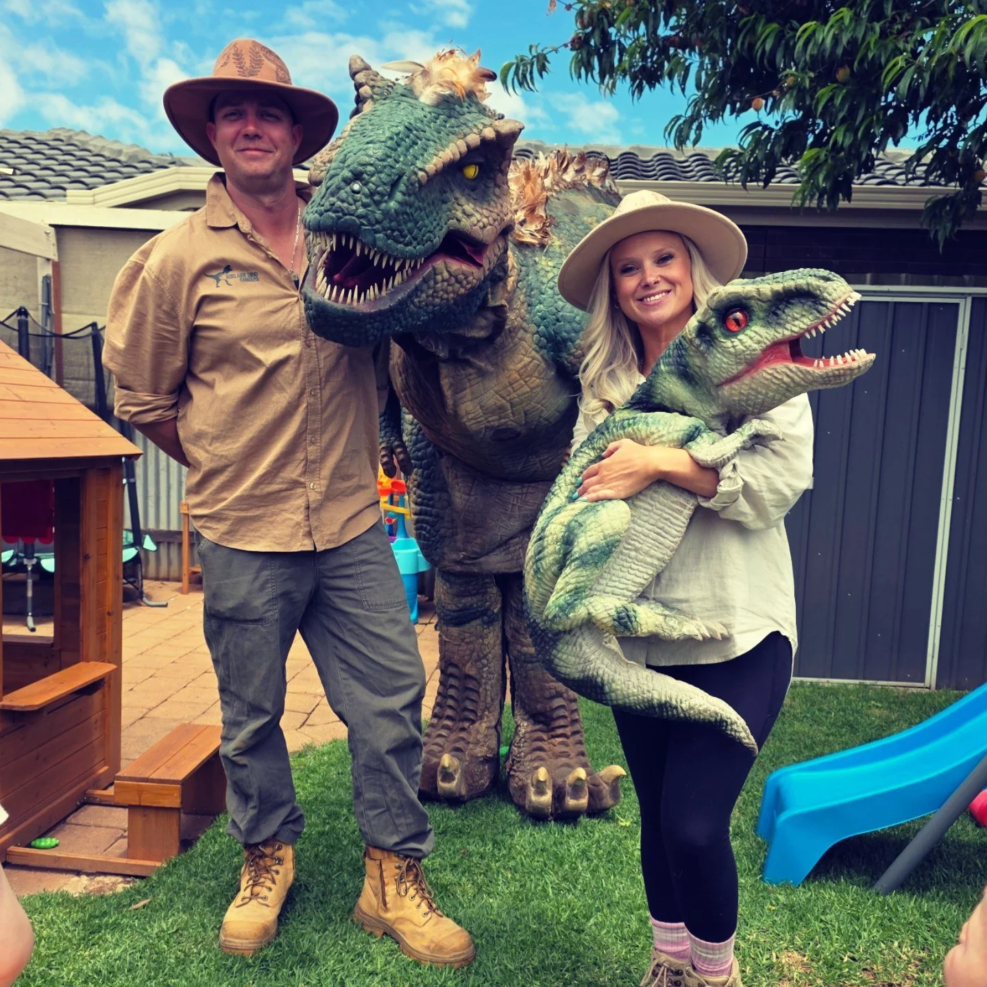 A man and a woman stand outdoors with two large dinosaur puppets, one resembles a Tyrannosaurus rex and the other a Velociraptor, during daytime.