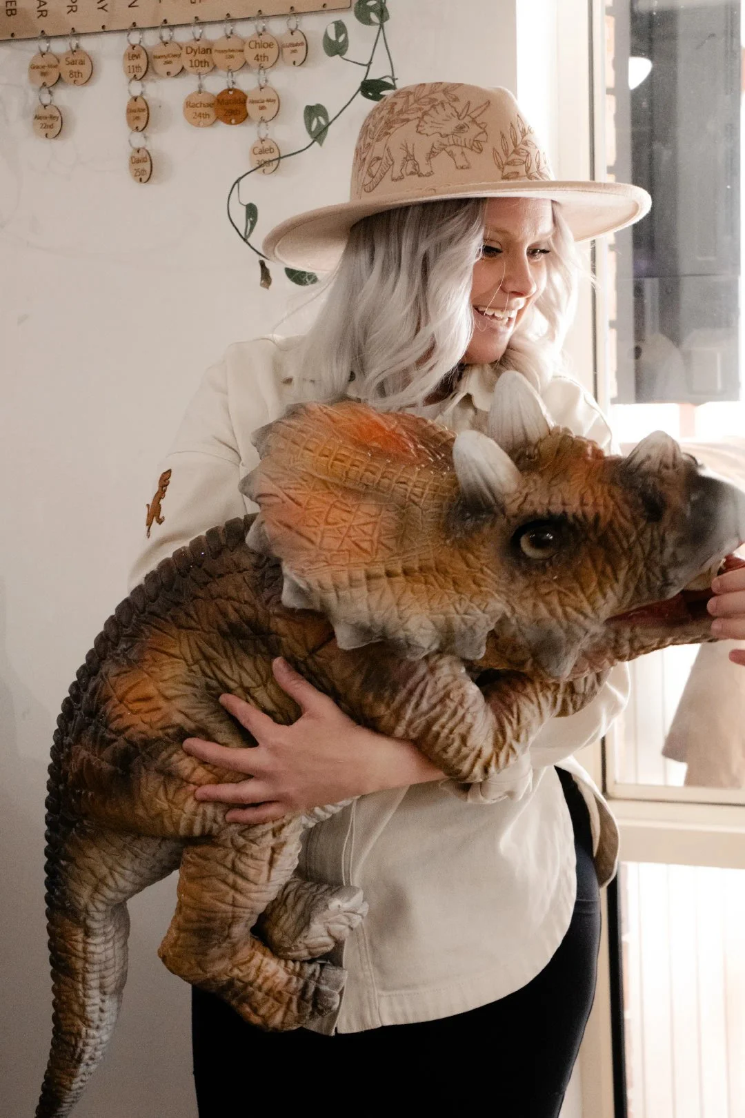 A woman with gray hair wearing a beige hat and shirt, holding a realistic-looking plush dinosaur toy in an indoor setting.