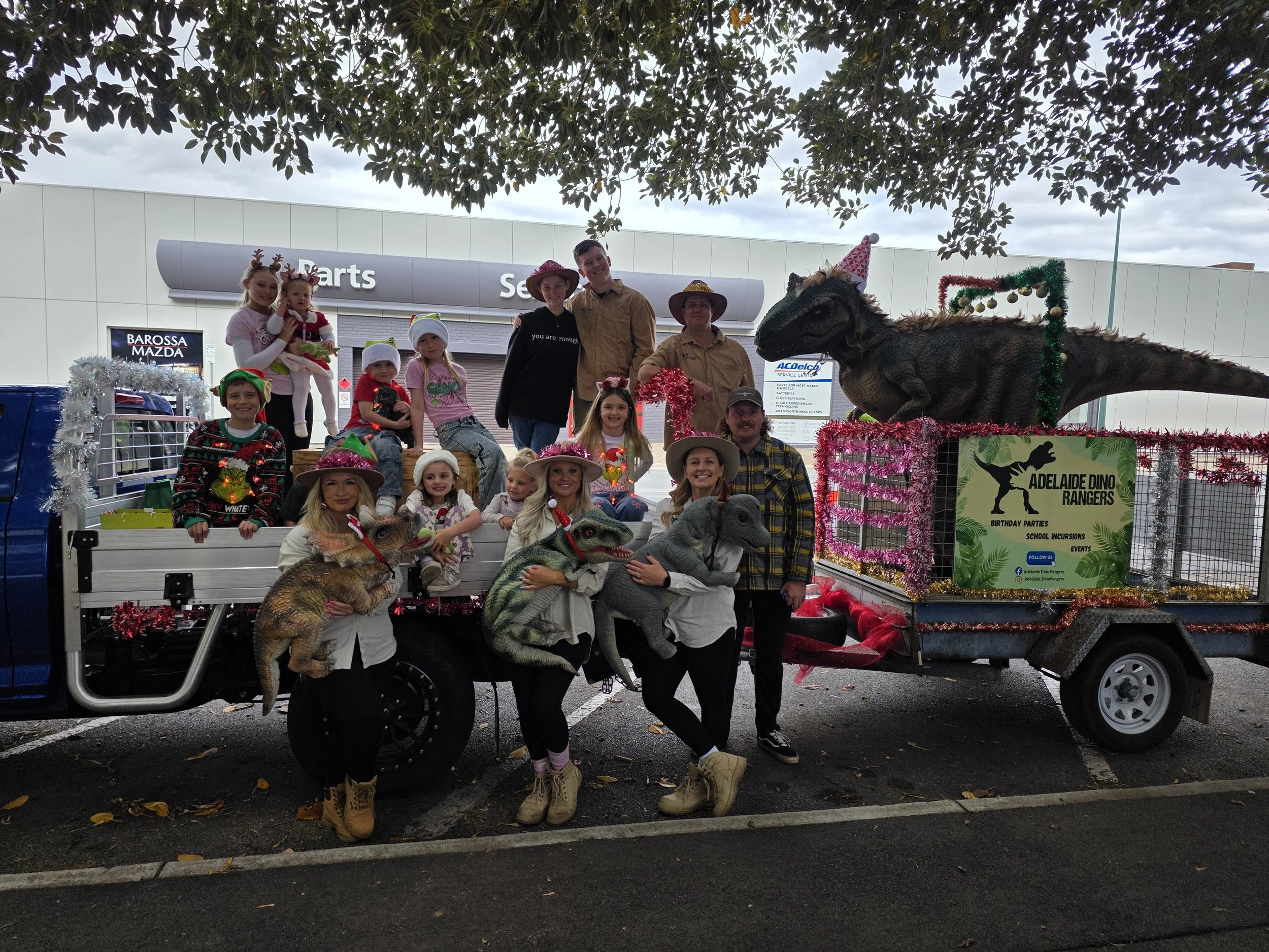 Group of children and adults on a parade float decorated with holiday tinsel, featuring dinosaur puppets and large dinosaur models, celebrating Christmas.