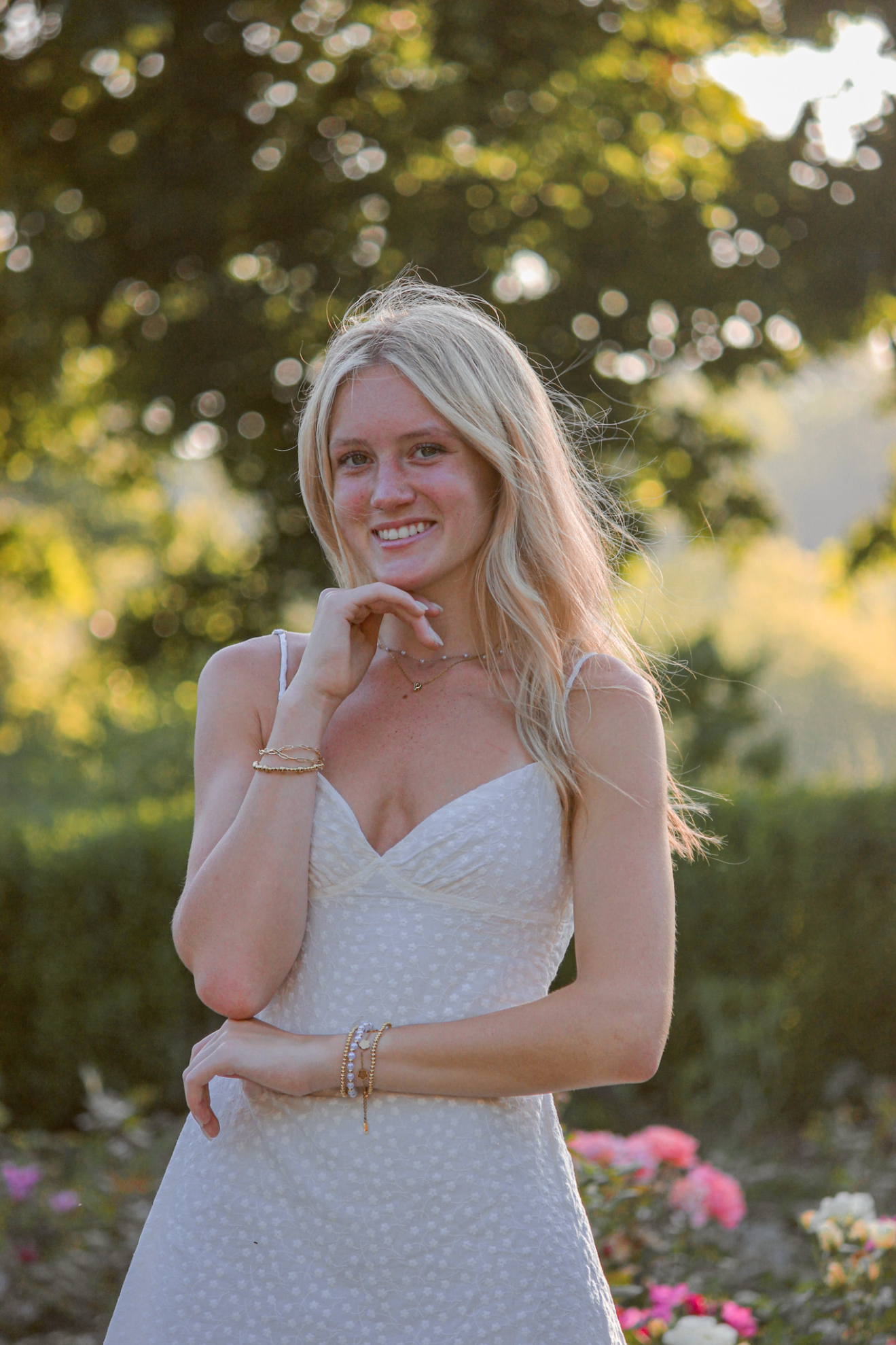 A young woman with long blonde hair in a white dress outdoors, smiling and posing with one hand near her face, surrounded by greenery and flowers.