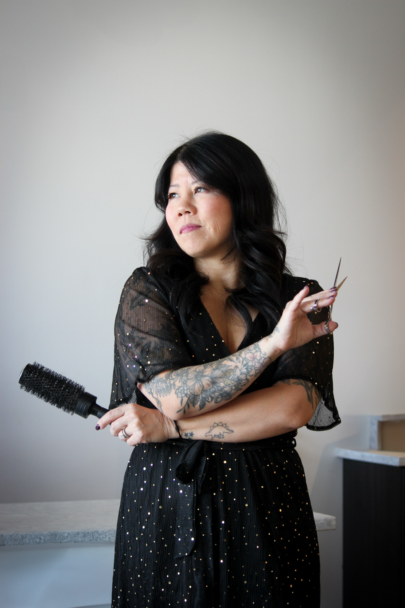 Woman with black hair and tattoos holding a round hairbrush and a pair of scissors, wearing a black dress with golden polka dots, standing against a plain light-colored wall.